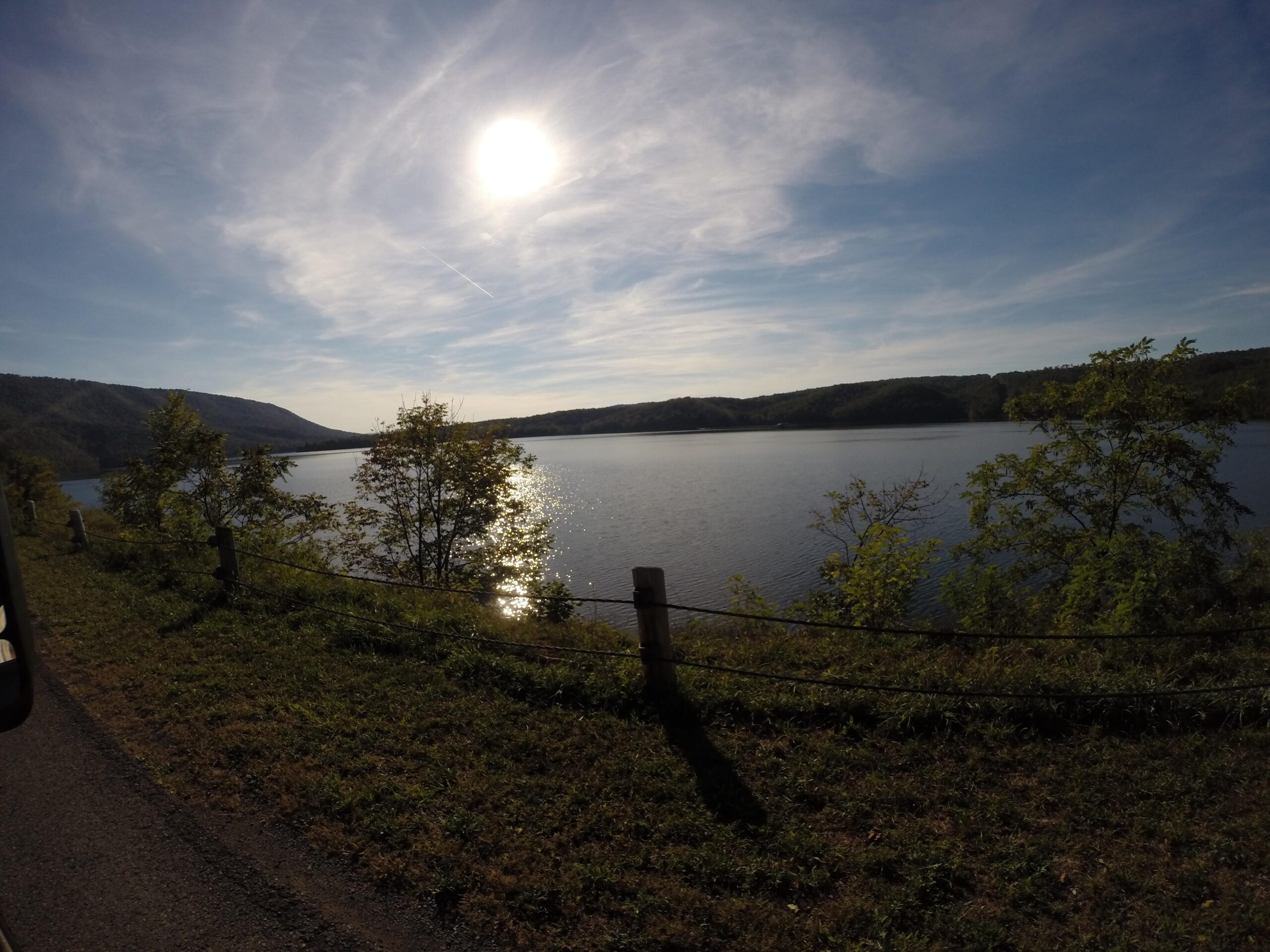 A serene landscape featuring a calm lake reflecting sunlight, surrounded by lush greenery and rolling hills under a blue sky with wispy clouds. A grassy area and wooden fence line the shore, creating a peaceful atmosphere. Allegrippis Trails mountain bike trail.
