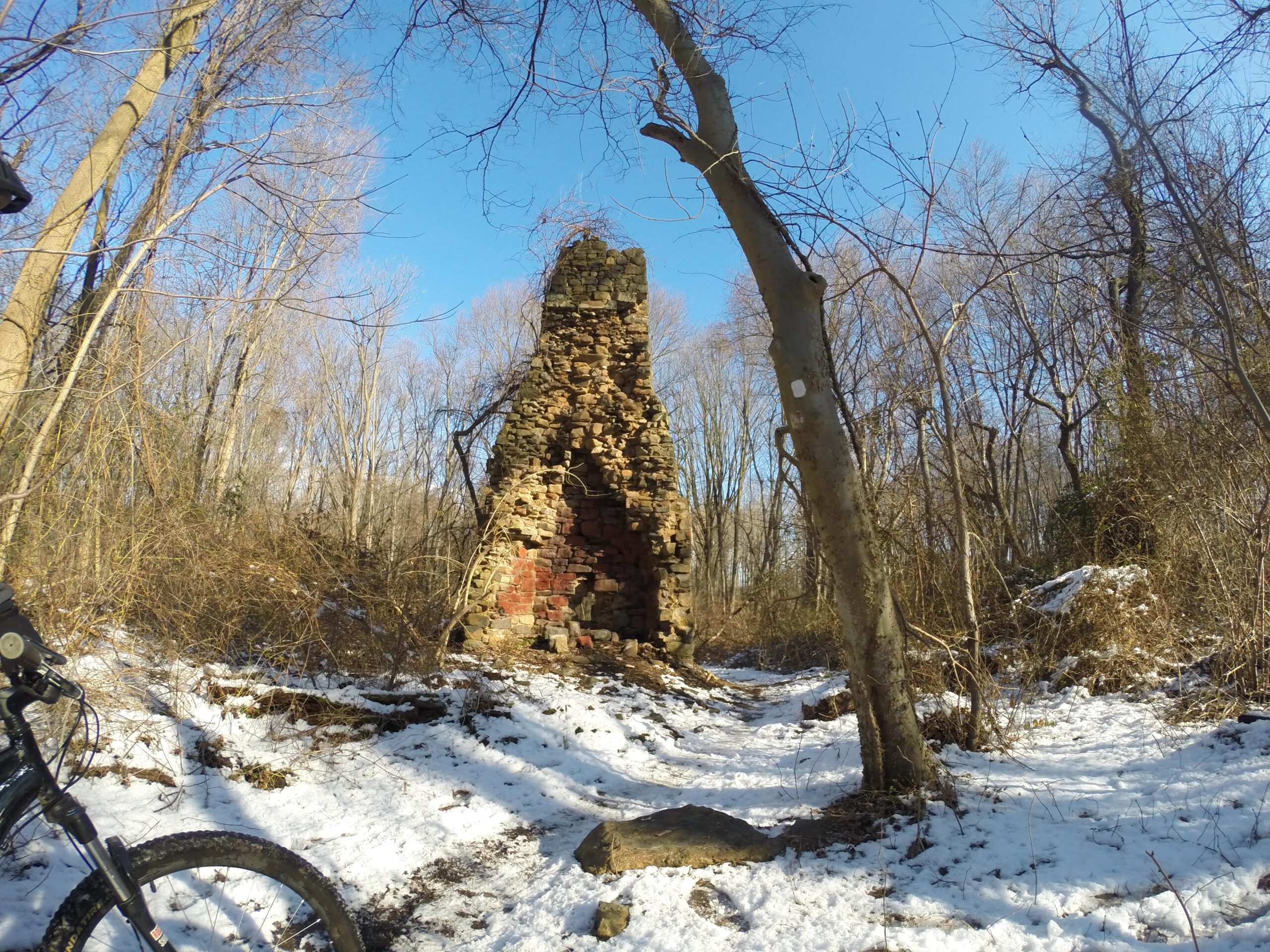 A stone chimney partially covered in snow stands amidst a forest with bare trees under a clear blue sky. The path leading to the chimney is lined with snow, while surrounding vegetation is sparse, indicating early spring or late winter conditions. A bicycle is partially visible in the foreground. Richmond Avenue and Forest Hill road mountain bike trail.