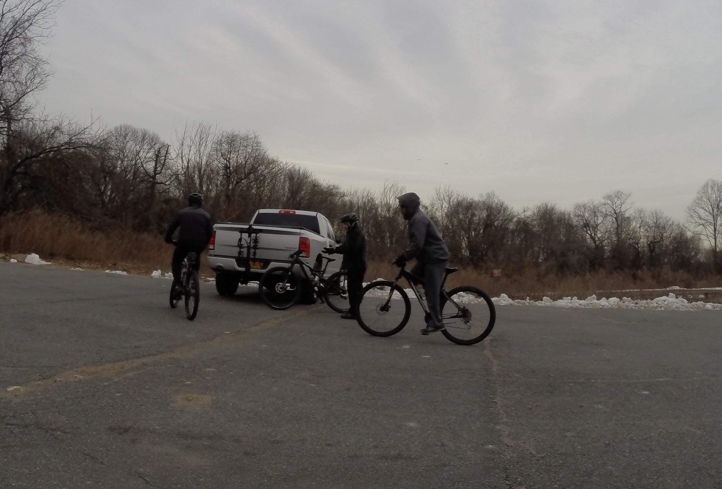 Three individuals riding bicycles in a parking area, with a white pickup truck parked nearby. The landscape is wooded with bare trees and some patches of snow on the ground. The sky is overcast, giving the scene a cool atmosphere. Richmond Avenue and Forest Hill road mountain bike trail.