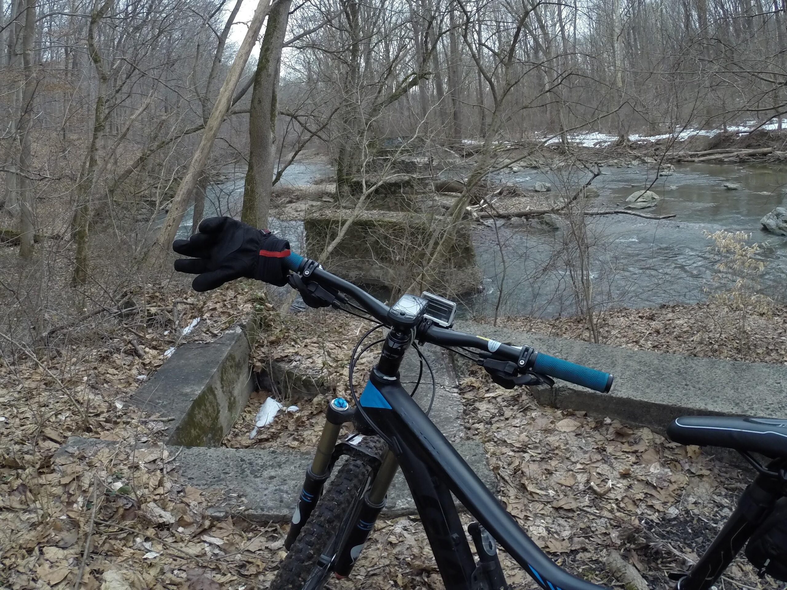 A mountain bike positioned on a trail next to a flowing creek, with bare trees and scattered leaves in the background. A gloved hand is raised in the foreground, indicating a pause or wave. Snow patches are visible along the edges of the creek. White Clay Creek mountain bike trail.
