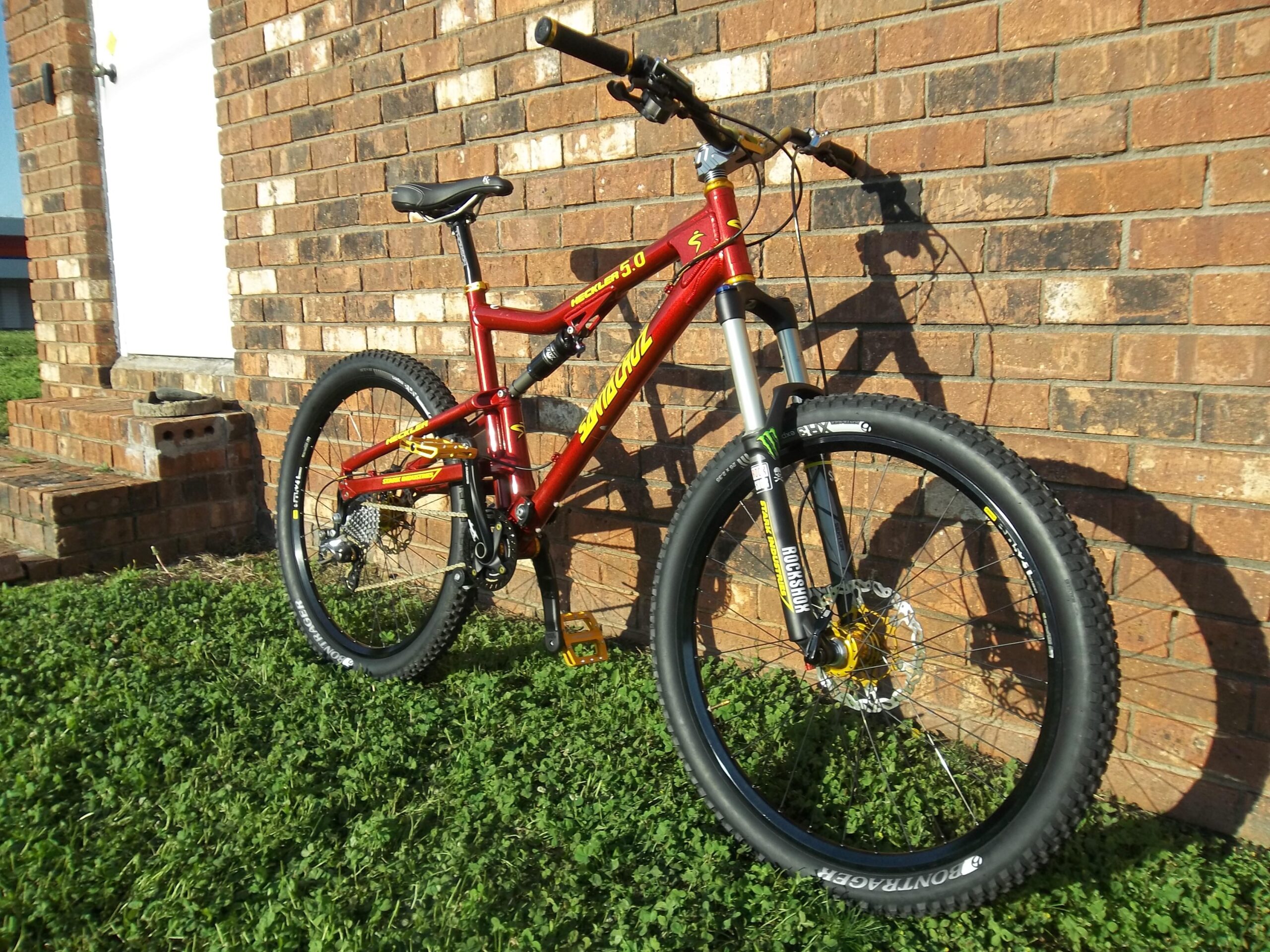 Santa Cruz Heckler 5.0: A red and yellow mountain bike is leaning against a brick wall, with green grass in the foreground. The bike features thick tires and a front suspension fork, showcasing its off-road capabilities. In the background, a white door and brick steps are visible.
