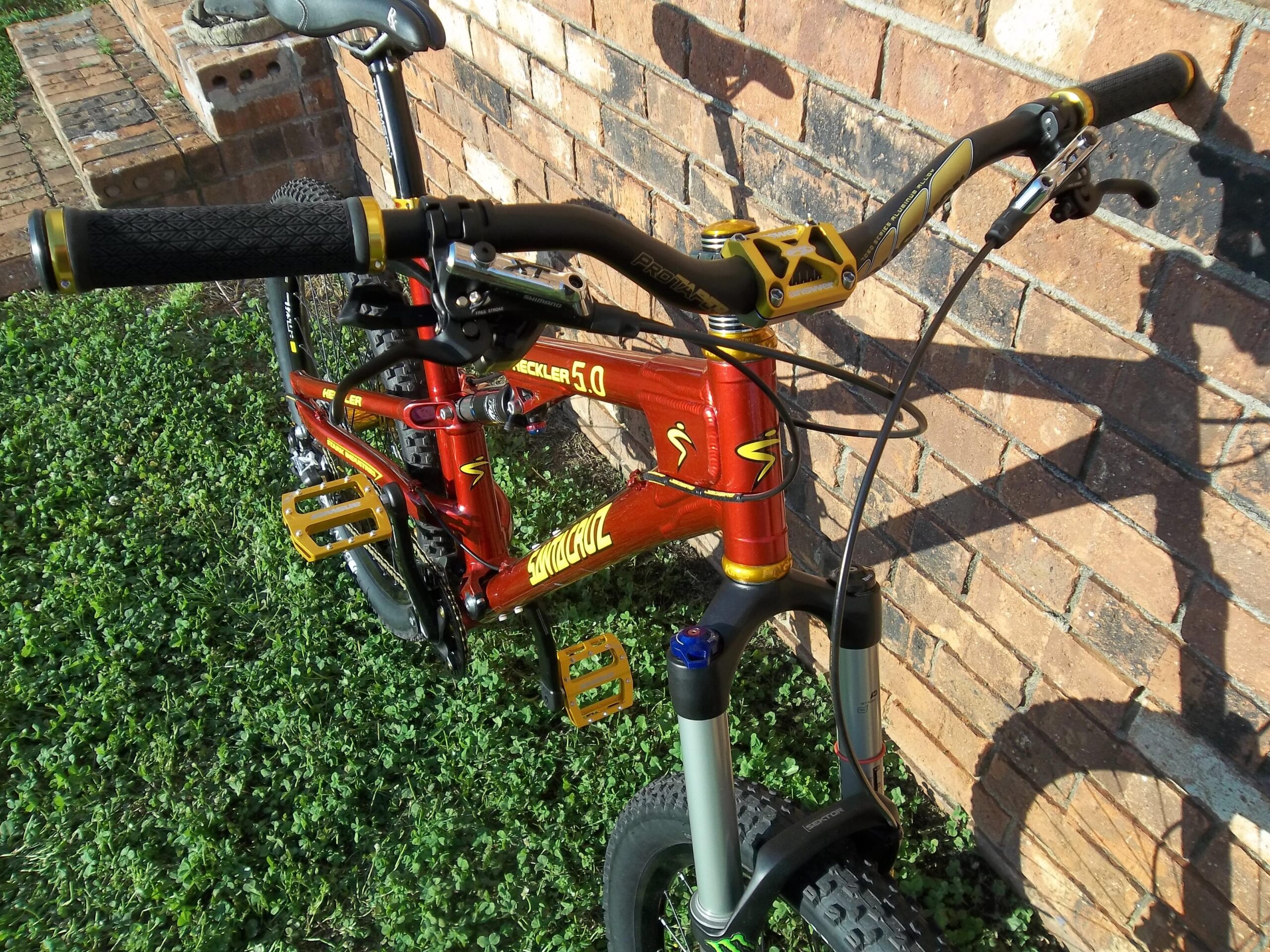 Santa Cruz Heckler 5.0: A close-up view of a red mountain bike leaning against a brick wall, featuring black handlebars with yellow accents, textured black grips, and gold pedals. The bike is positioned on green grass, showcasing its front suspension and prominent branding details.