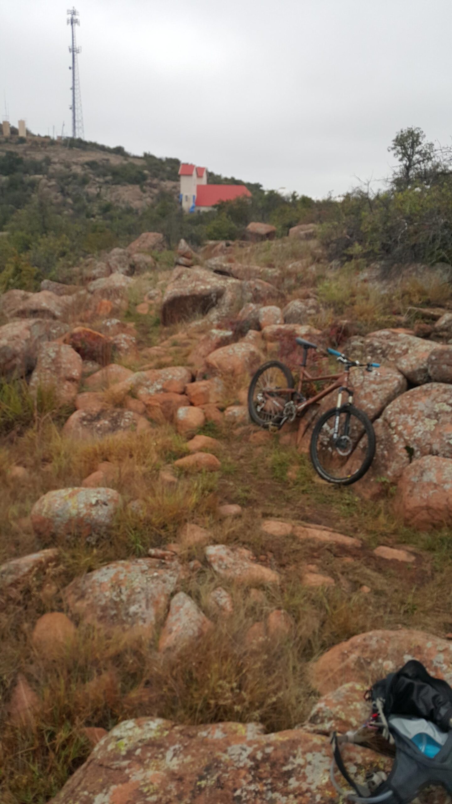 A mountain bike resting on a rocky trail surrounded by grass and small bushes, with a hillside and a small building in the background. The sky is overcast, suggesting a cloudy day. Lake Lawtonka Trails mountain bike trail.