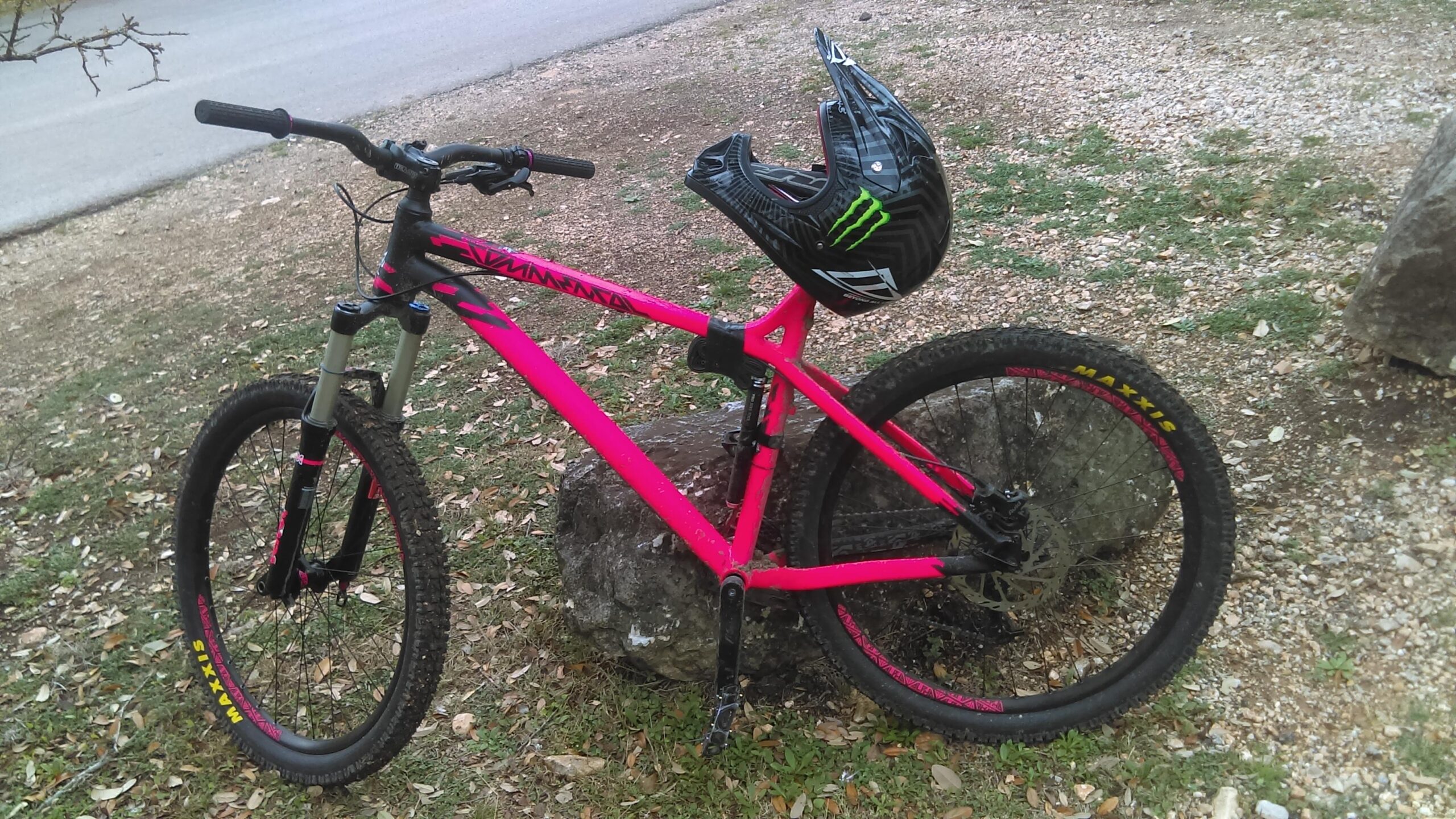 Commencal Meta HT 1: A bright pink mountain bike leaning against a large rock, with a black helmet resting on the handlebars. The background shows a grassy area with scattered leaves and a road in the distance. The bike features thick tires and a sturdy frame, designed for off-road riding.