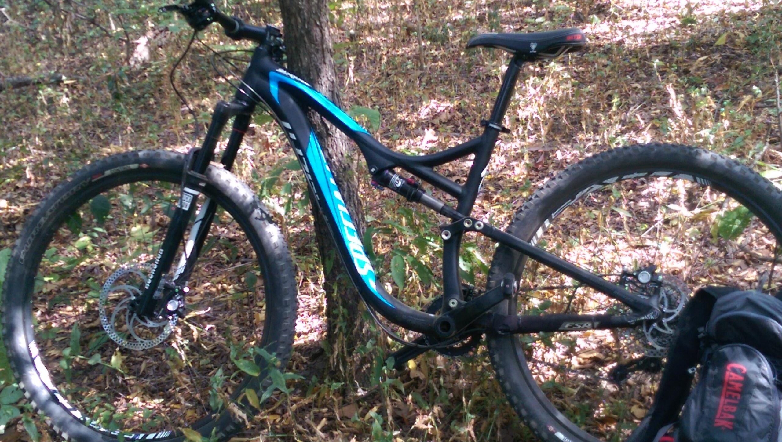 Specialized Stumpjumper FSR Comp 29er: A mountain bike resting on the ground amidst fallen leaves and greenery in a wooded area. The bike features a sleek black frame with blue accents and large, knobby tires suitable for off-road terrain. A CamelBak bag is visible in the foreground, suggesting a biking adventure.