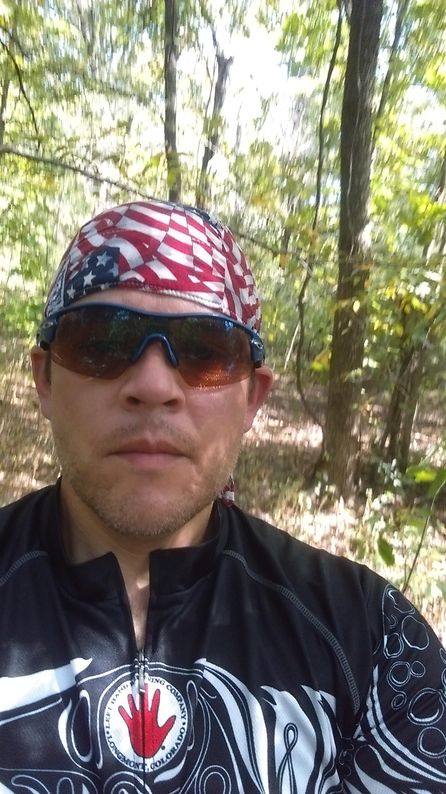 Specialized Stumpjumper FSR Comp 29er: A person wearing sunglasses and a bandana featuring an American flag design, standing in a wooded area with trees and foliage in the background. The individual is dressed in a black cycling jersey with a logo on the front.