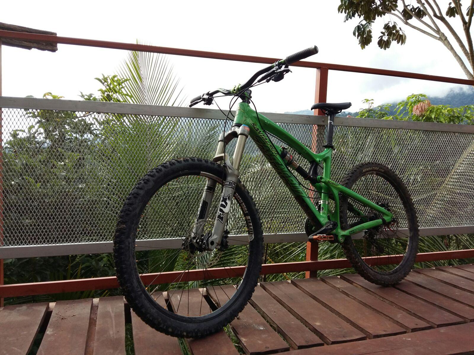Santa Cruz Nomad: A green mountain bike parked on a wooden deck, with a mesh railing and a lush green landscape in the background, including trees and distant mountains under a cloudy sky.