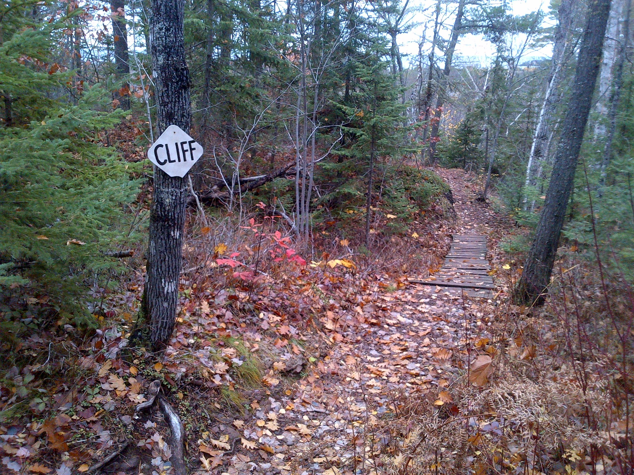 A forested pathway with colorful autumn leaves, featuring a warning sign that reads "CLIFF" attached to a tree on the left. The path is narrow, winding through trees, with some wooden planks visible, indicating it may lead to an overlook. Copper Harbor Trails mountain bike trail.