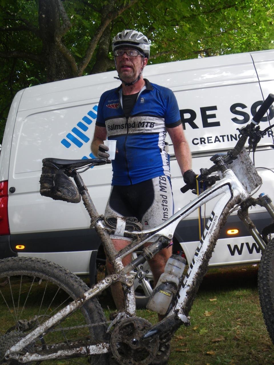 Specialized Stumpjumper FSR: A mountain biker, covered in mud, stands beside a dirty bicycle in a wooded area. He wears a blue cycling jersey with "Halmstad MTB" printed on it and has protective gear, including glasses and a helmet. The background features a delivery van with a partially obscured logo. The cyclist holds a cup and looks tired yet satisfied after a challenging ride.