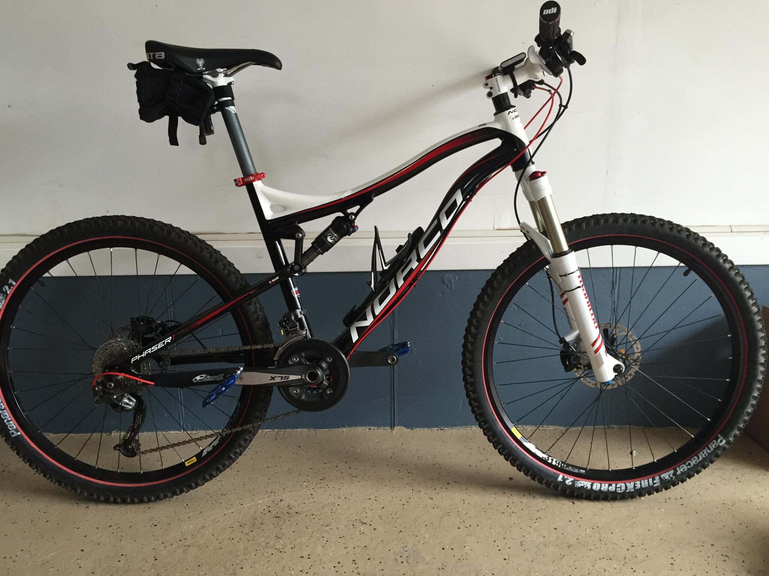 Norco Phaser 1: A black and white mountain bike with red accents, featuring a raised seat, front suspension, and knobby tires. The bike is positioned against a wall with a blue lower section and a light-colored upper section, on a speckled floor. A small black saddle bag is attached to the seat post.