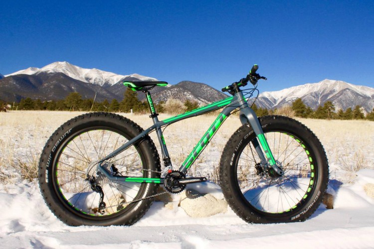 A green and gray fat bike positioned on a snowy landscape, with majestic snow-capped mountains in the background and a clear blue sky overhead. The bike features wide tires designed for optimal traction on snow and terrain.