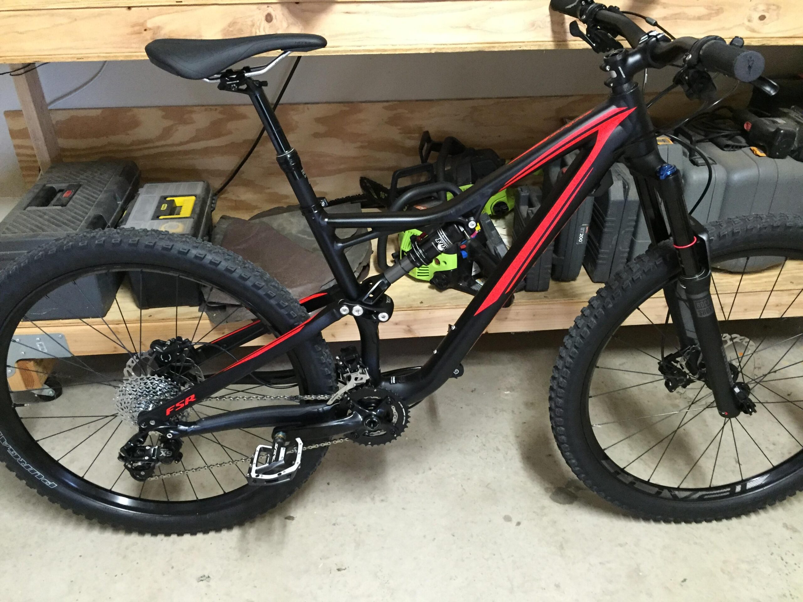 Specialized Stumpjumper FSR: A black and red mountain bike is resting on a concrete floor, with a wooden shelf in the background holding various tools and equipment. The bike features thick tires, a prominent suspension system, and a modern frame design. The setting appears to be a garage or workshop.