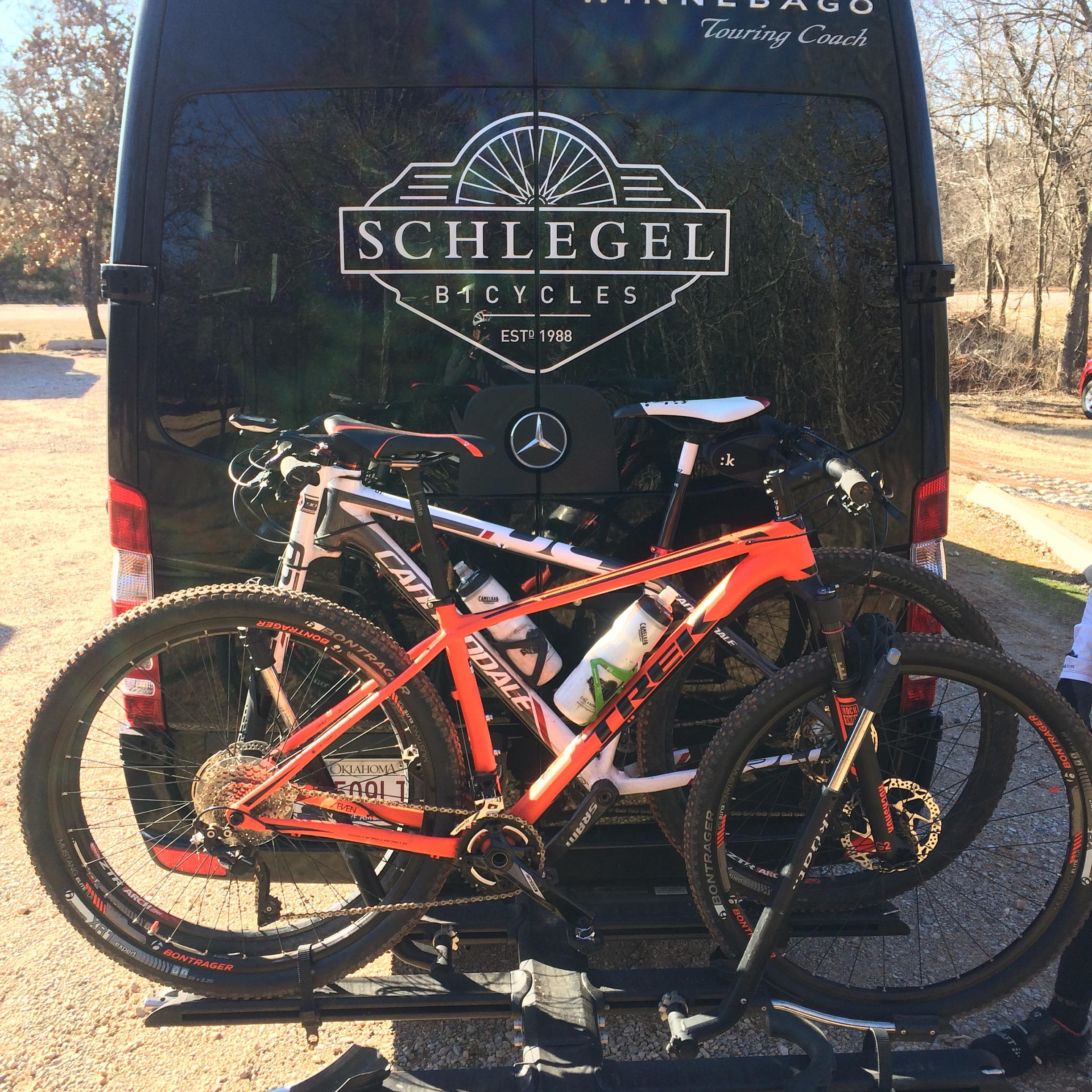 Trek Superfly 7: Image of a black touring van with the logo "Schlegel Bicycles" and "EST 1988" on its back. Two mountain bikes are securely mounted on a bike rack attached to the rear. One bike is orange, a Trek model, while the other is white, a Cannondale model. The setting appears outdoor, with trees visible in the background.