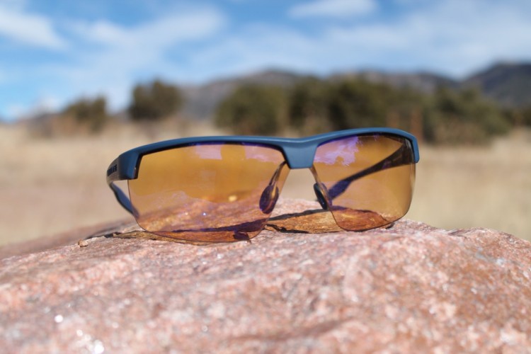 A pair of sunglasses with tinted lenses resting on a rock, set against a backdrop of a grassy field and blue sky with scattered clouds.