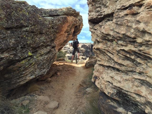 A mountain biker navigating a narrow path between two large rock formations in a rugged outdoor setting. The trail is sandy and leads through a rocky landscape under a partly cloudy sky. Zen Trail mountain bike trail.