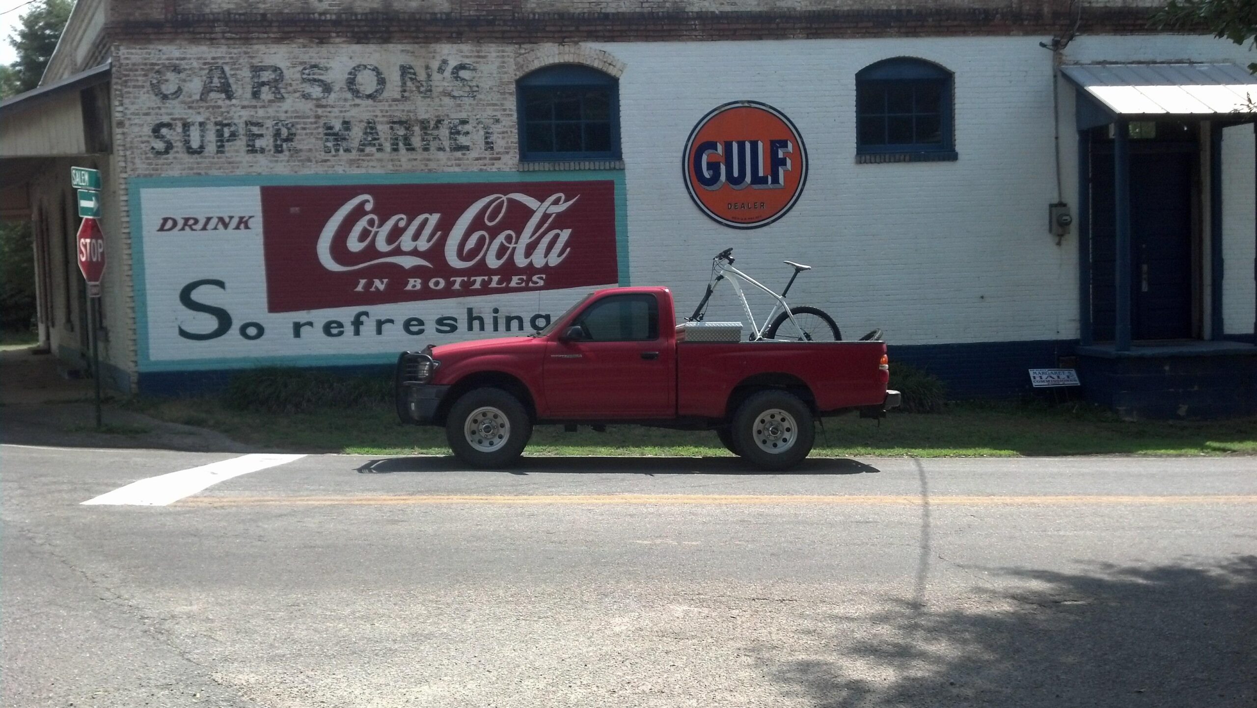 A red pickup truck parked on the side of the road in front of a vintage building. The building features a large sign that reads "CARSON'S SUPER MARKET" and advertisements for Coca-Cola. A white bicycle is secured in the truck's bed, and a stop sign is visible in the foreground. The scene captures a quaint, small-town atmosphere. Heritage Park mountain bike trail.