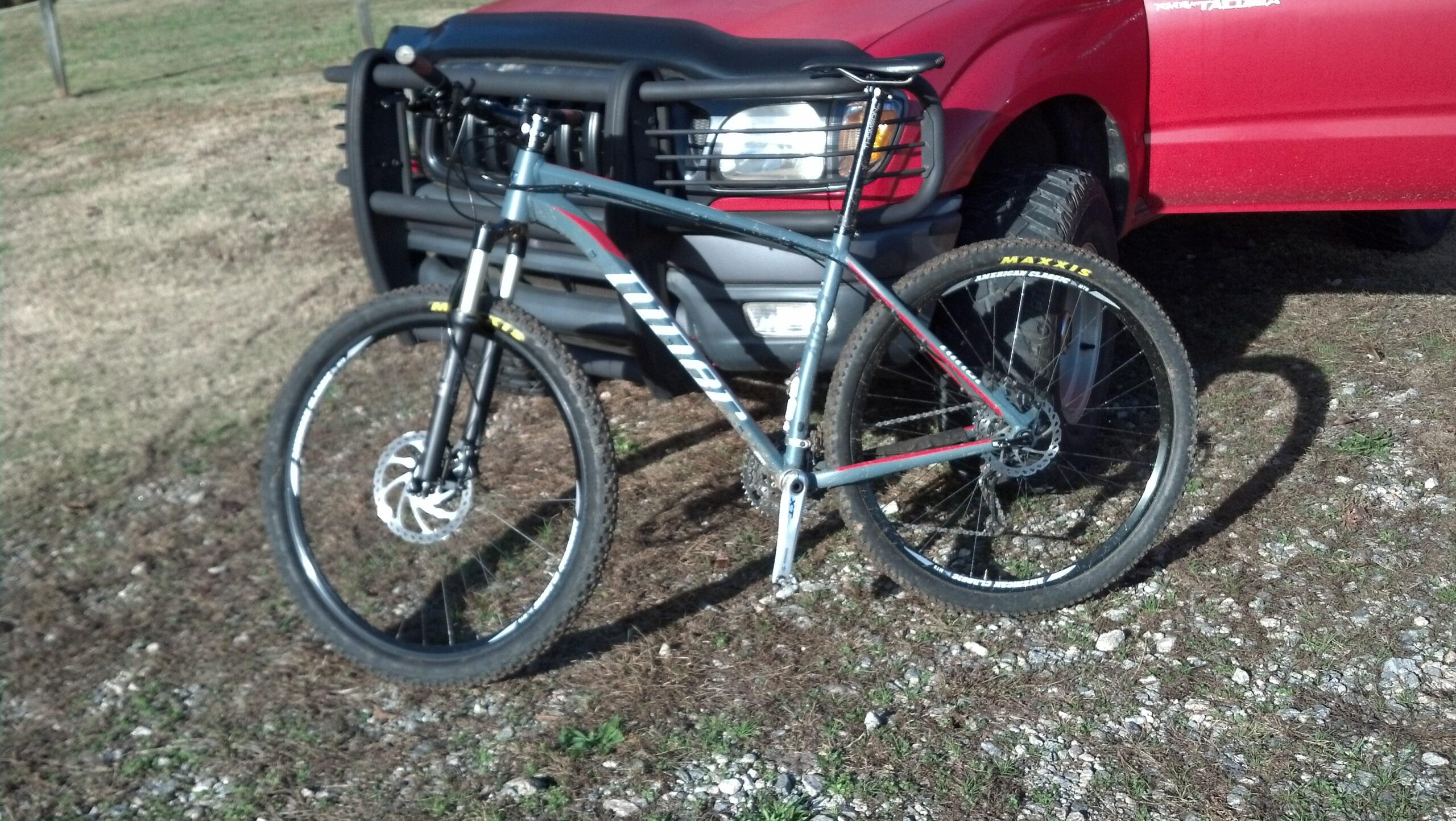 Niner Air 9: A mountain bike with a gray frame and black forks is leaning against a red pickup truck. The bike features large tires with "Maxxis" branding and a disc brake system. The background consists of a grassy field with some gravel visible.