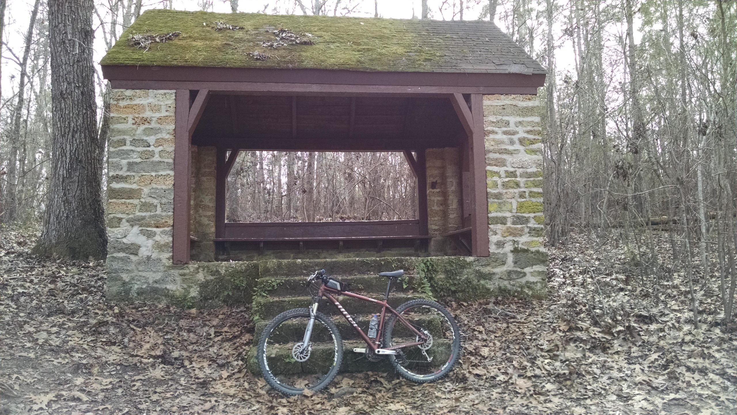 A rustic stone pavilion covered with moss, surrounded by bare trees in a wooded area. In the foreground, a mountain bike rests against the steps leading up to the pavilion, which features a wooden frame and an open front. Leaves are scattered on the ground. The Knot mountain bike trail.