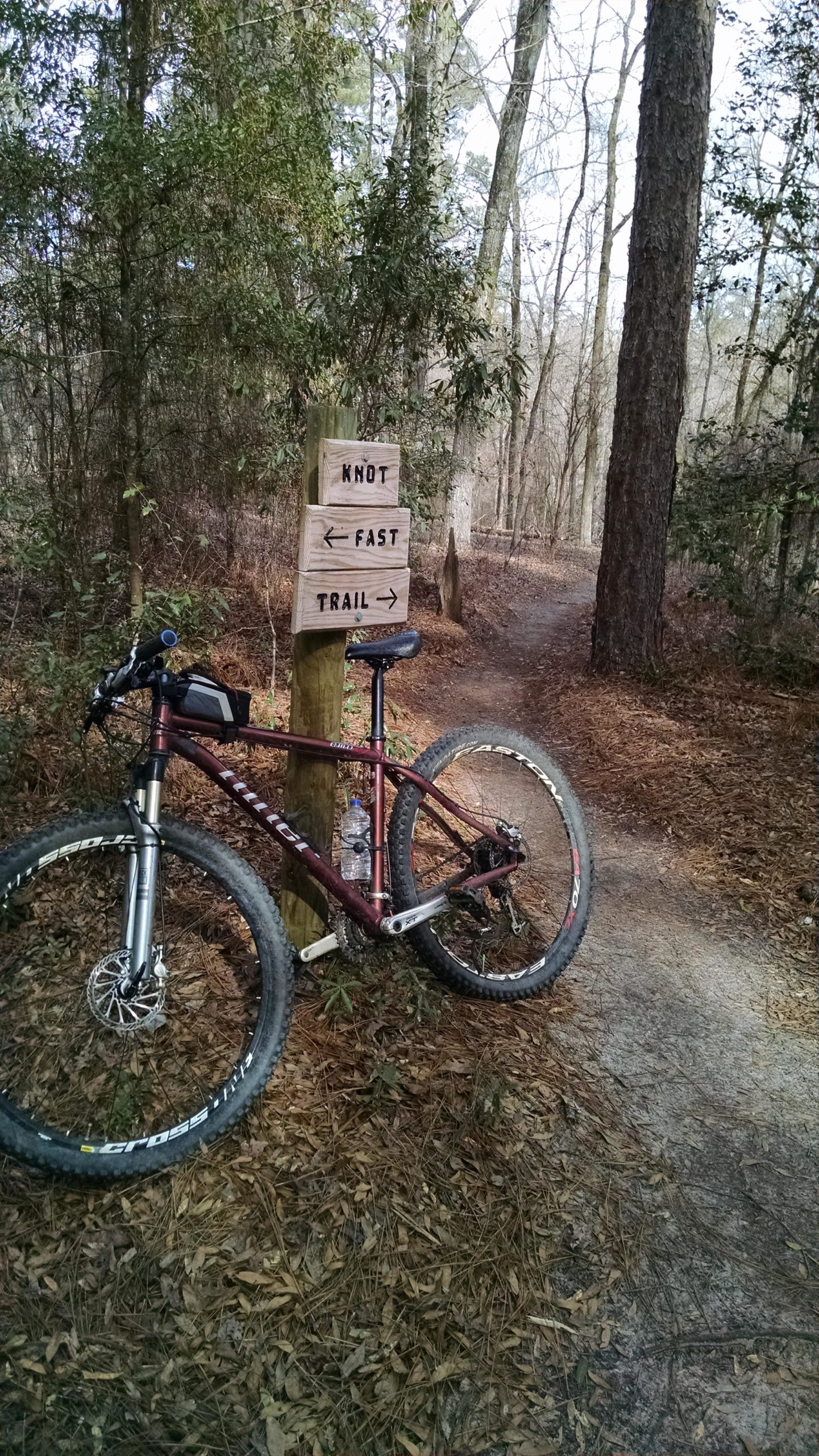 A mountain bike leaning against a wooden trail sign, which indicates directions for the "Knot" and "Fast" trails. The background features a forested area with trees and fallen leaves, and a winding dirt path leading into the woods. The Knot mountain bike trail.
