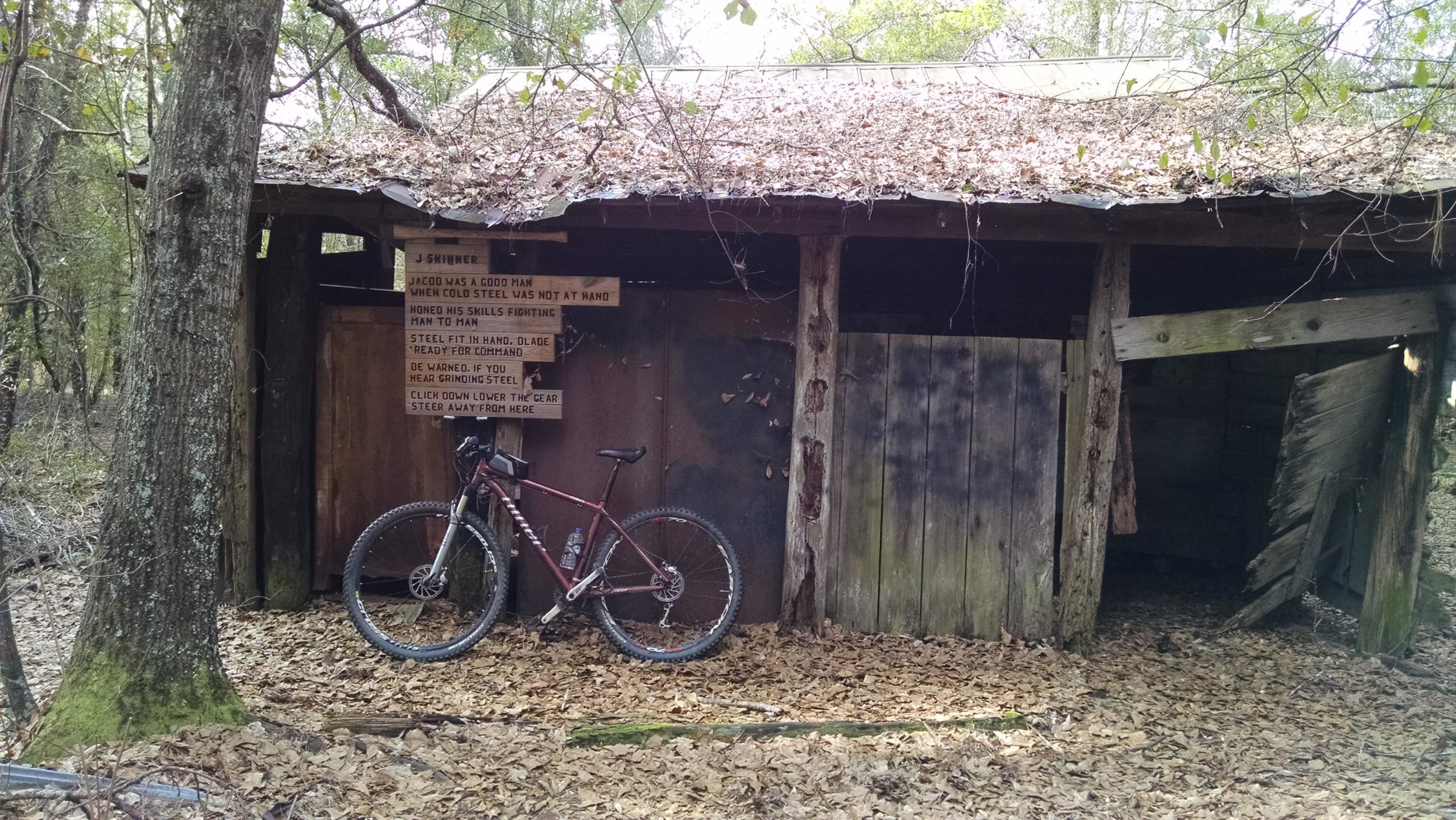 An old, weathered wooden shed with a roof covered in leaves, surrounded by trees. A mountain bike leans against the shed, and a wooden sign nearby features text honoring a man named Jacob Skinner. The ground is covered with fallen leaves. The Knot mountain bike trail.