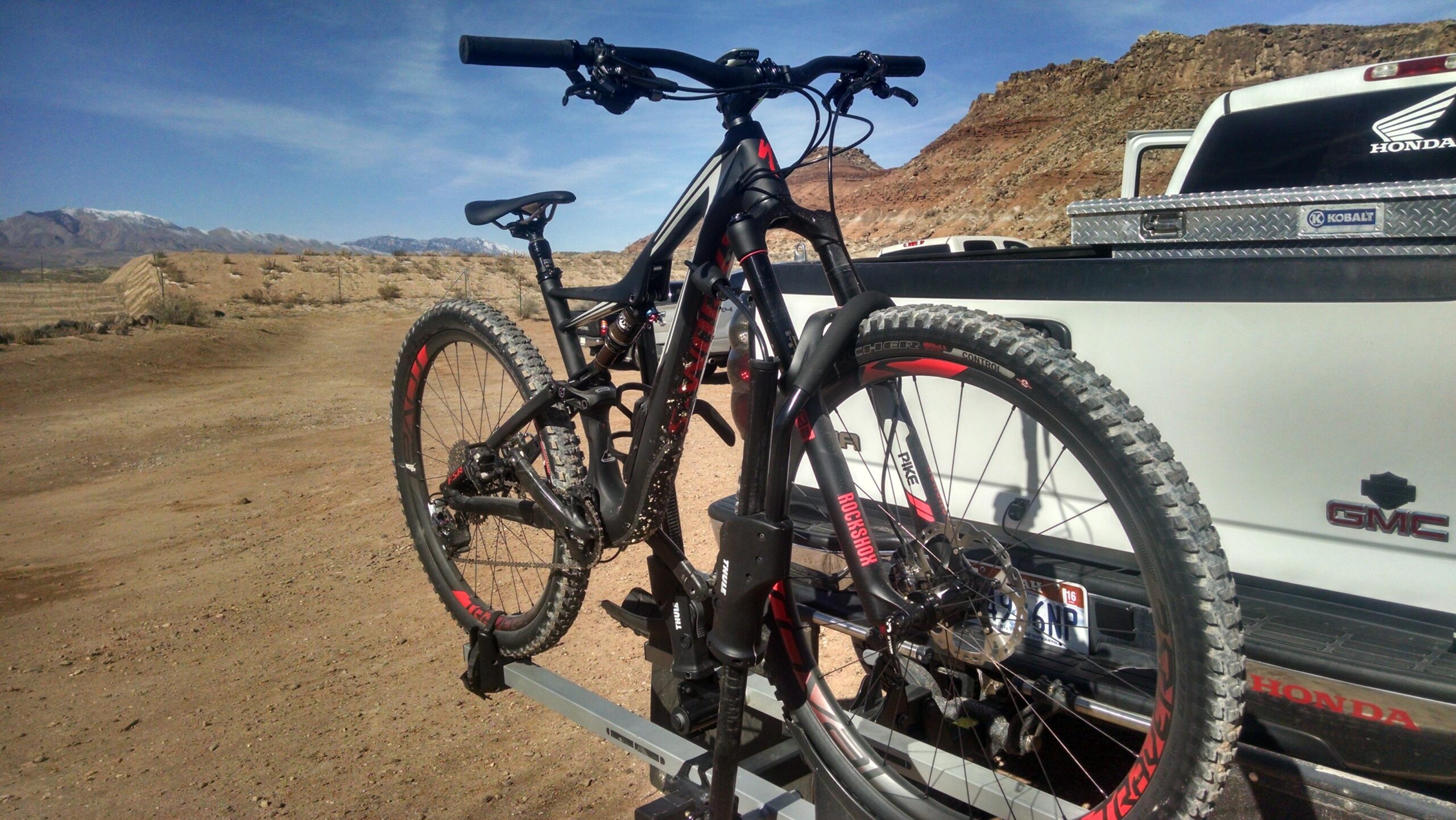 Specialized SWORKS Stumpjumper FSR: A close-up view of a mountain bike secured on a bike rack, with a pickup truck in the background. The bike features black and red accents, and the setting includes a mountain landscape under a clear blue sky. The dirt terrain suggests a rugged outdoor environment, ideal for biking activities.