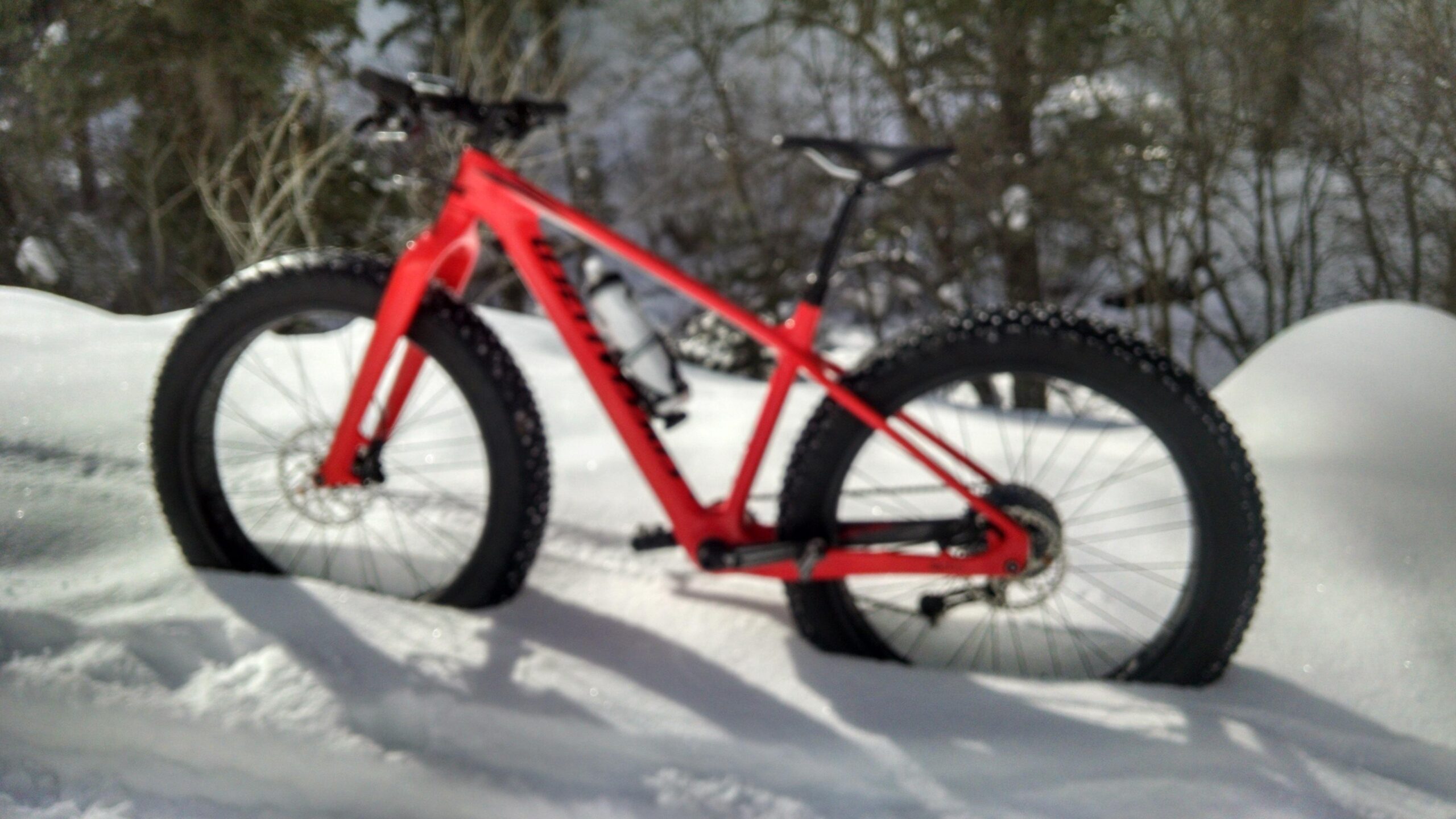 Specialized Fatboy Carbon Comp: A bright red fat tire bike resting on a snowy landscape, with large, wide tires designed for winter conditions. The bike is partially obscured by snow, and there are trees in the background, suggesting a wintery outdoor setting.