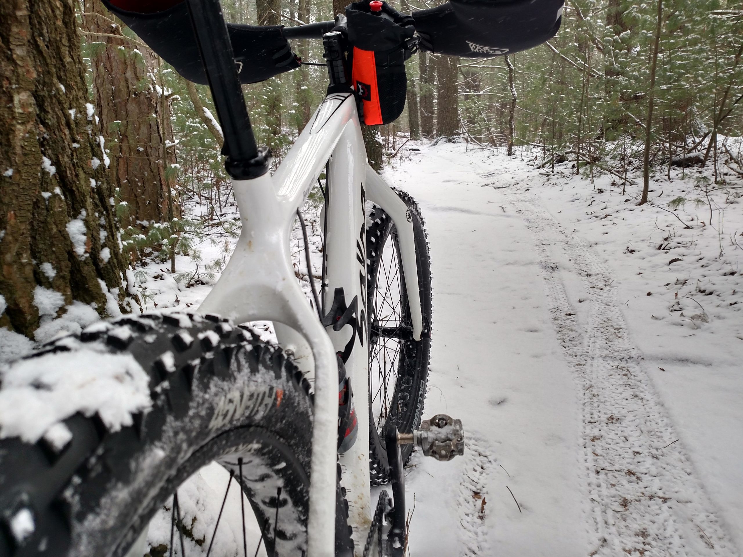 A close-up view of a white bike with thick tires, positioned on a snowy forest trail. Snow-covered trees are visible in the background, creating a wintery scene. The bike's front tire is prominently displayed, partially covered in snow. Yankee Springs mountain bike trail.