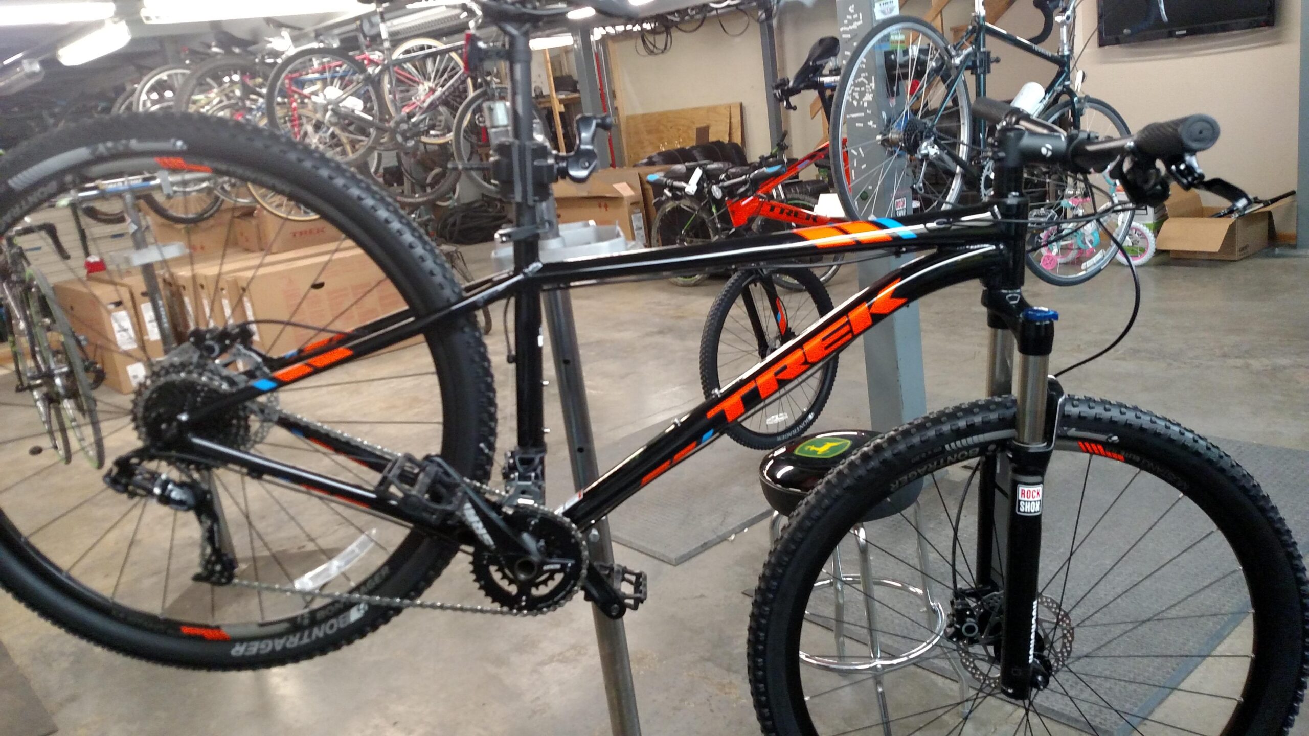 Trek X-Caliber: Alt text: A black Trek mountain bike is in a bike shop, mounted on a repair stand. The bike features orange accents, a front suspension fork, and a detailed rear derailleur. In the background, various other bicycles are visible, along with cardboard boxes.