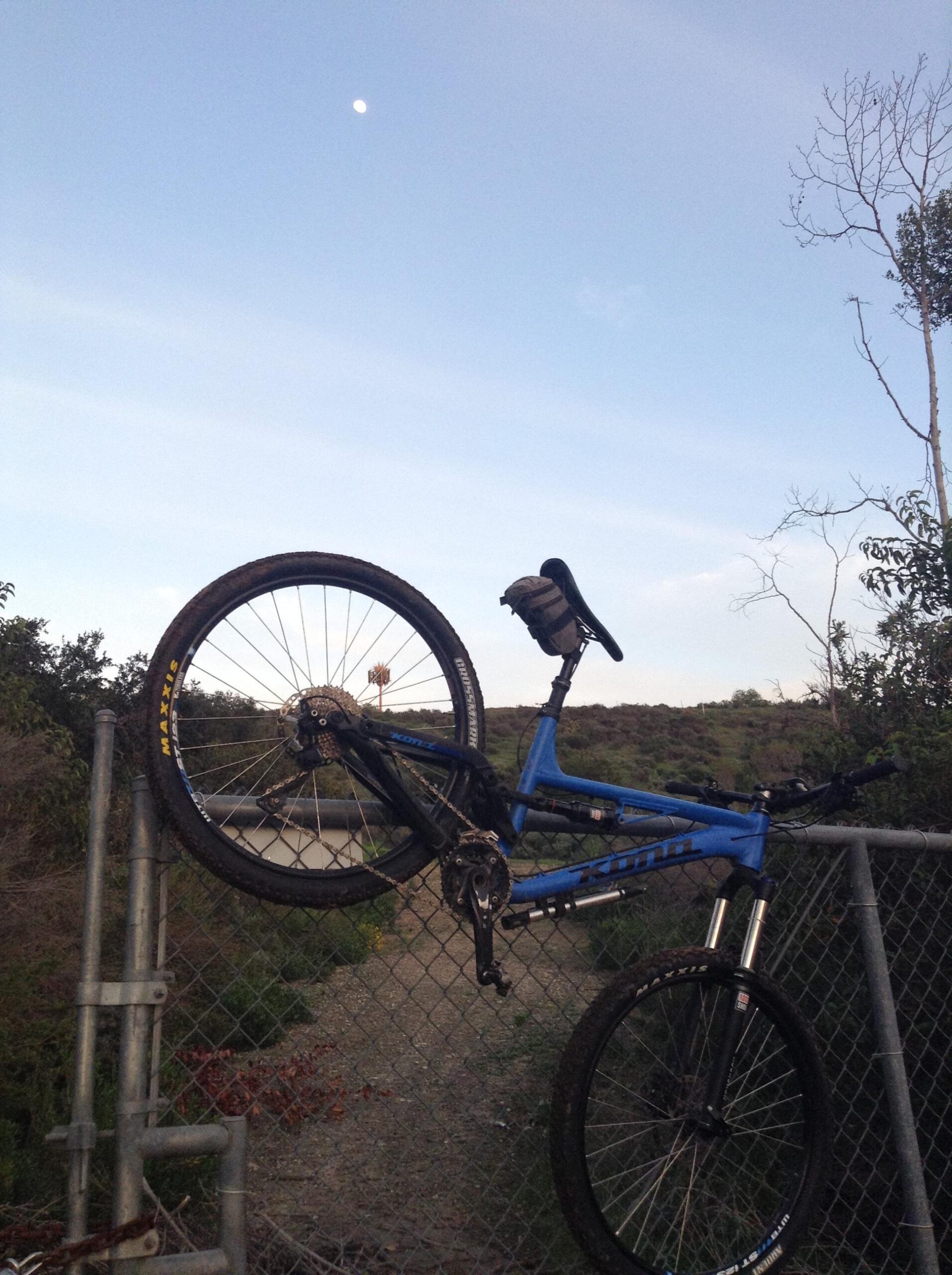 Kona Process 134: A blue mountain bike is resting on a chain-link fence with its rear wheel hanging over the edge. The sky is clear with a visible moon, and the background features a dirt path and sparse vegetation.