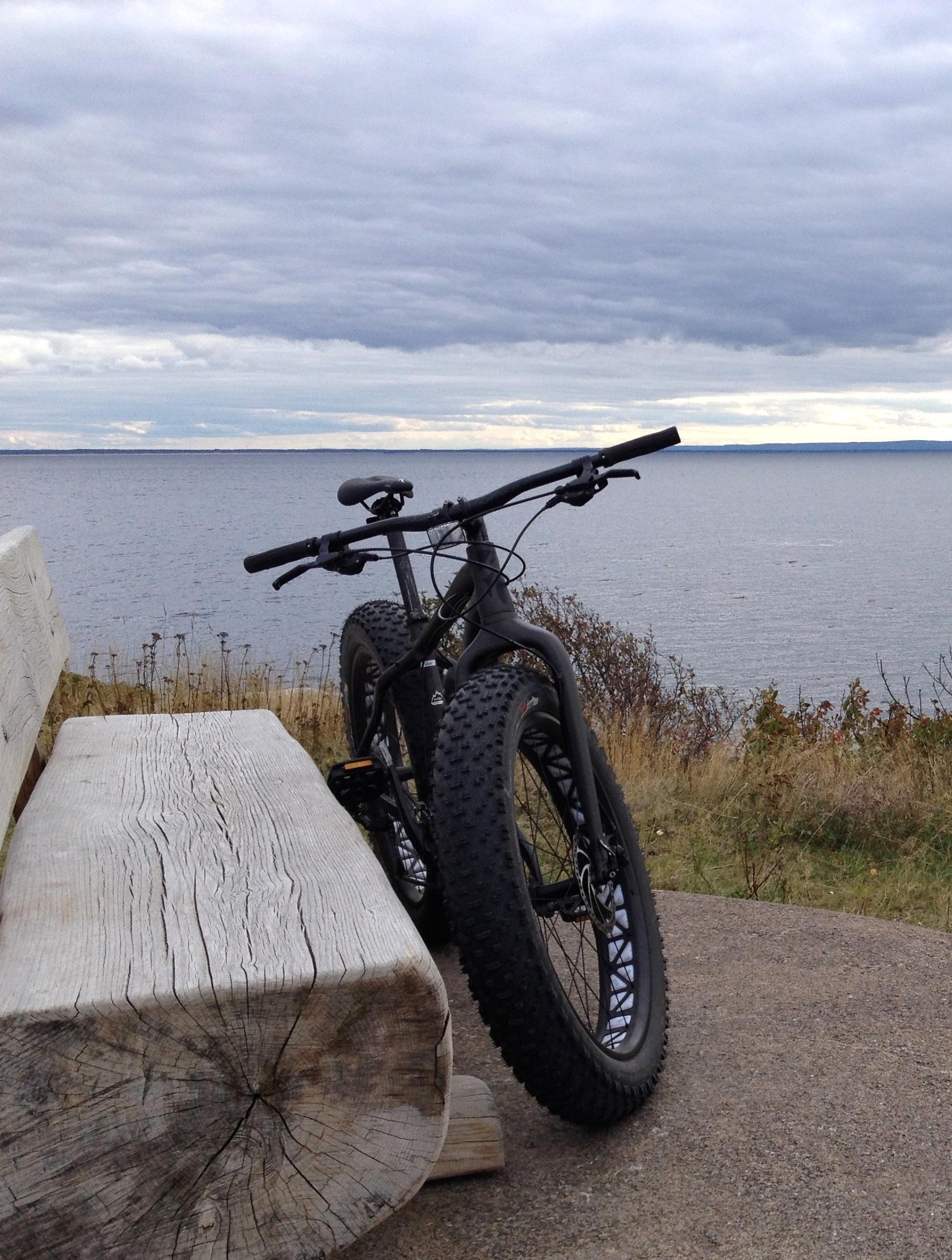 Specialized Fatboy: A black fat bike parked beside a weathered wooden bench, overlooking a calm lake with a cloudy sky in the background. The scene captures a tranquil outdoor setting, emphasizing nature and adventure.