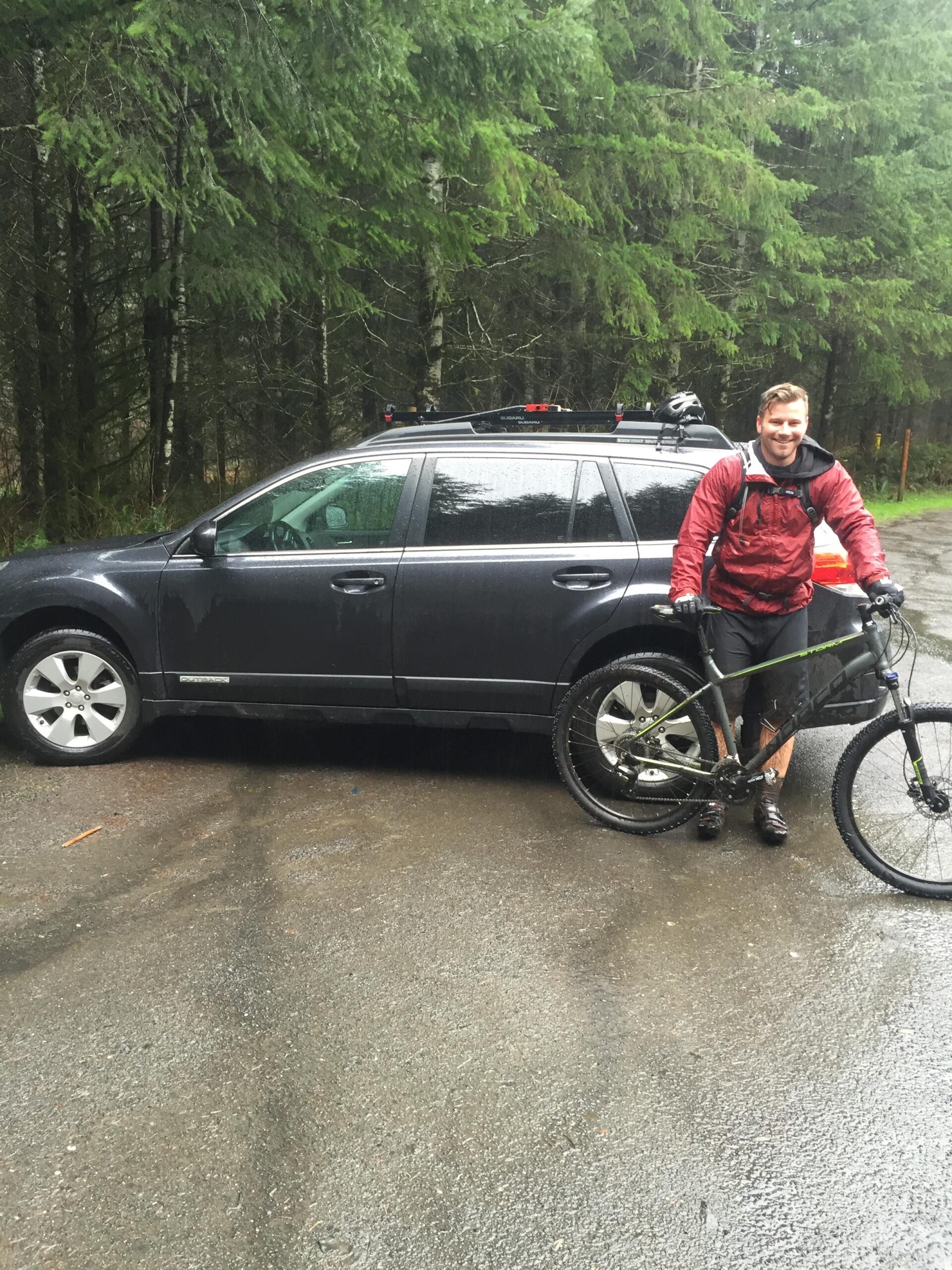 Norco Storm: A person wearing a red jacket and black shorts stands next to a parked car, holding a mountain bike. The background features lush green trees and a wet road, suggesting recent rain. The setting appears to be a forested area, ideal for outdoor activities.