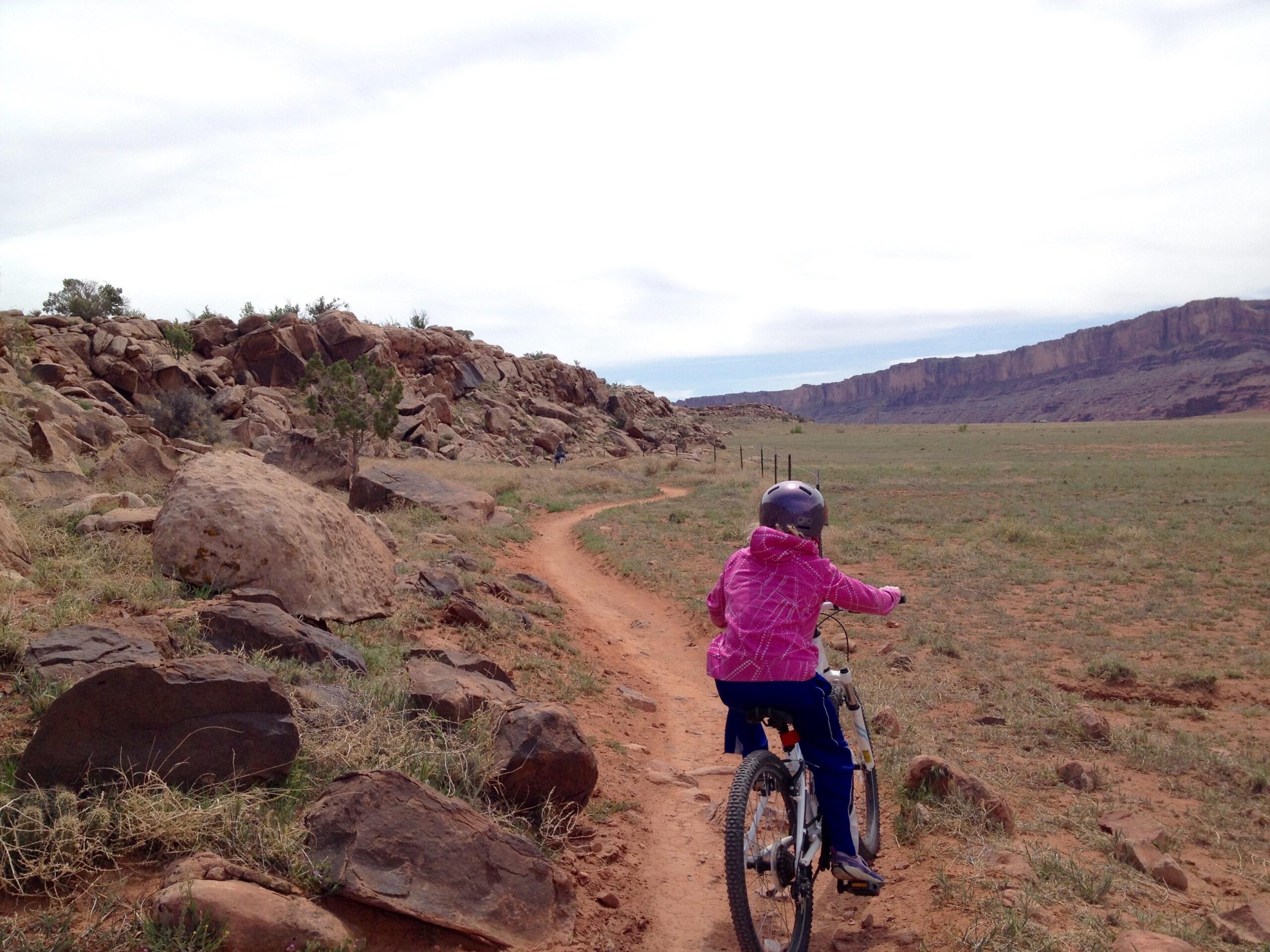 A child wearing a pink jacket and helmet rides a bicycle along a dirt path in a rocky landscape, with tall cliffs visible in the distance under a cloudy sky. Moab Brand Trails mountain bike trail.