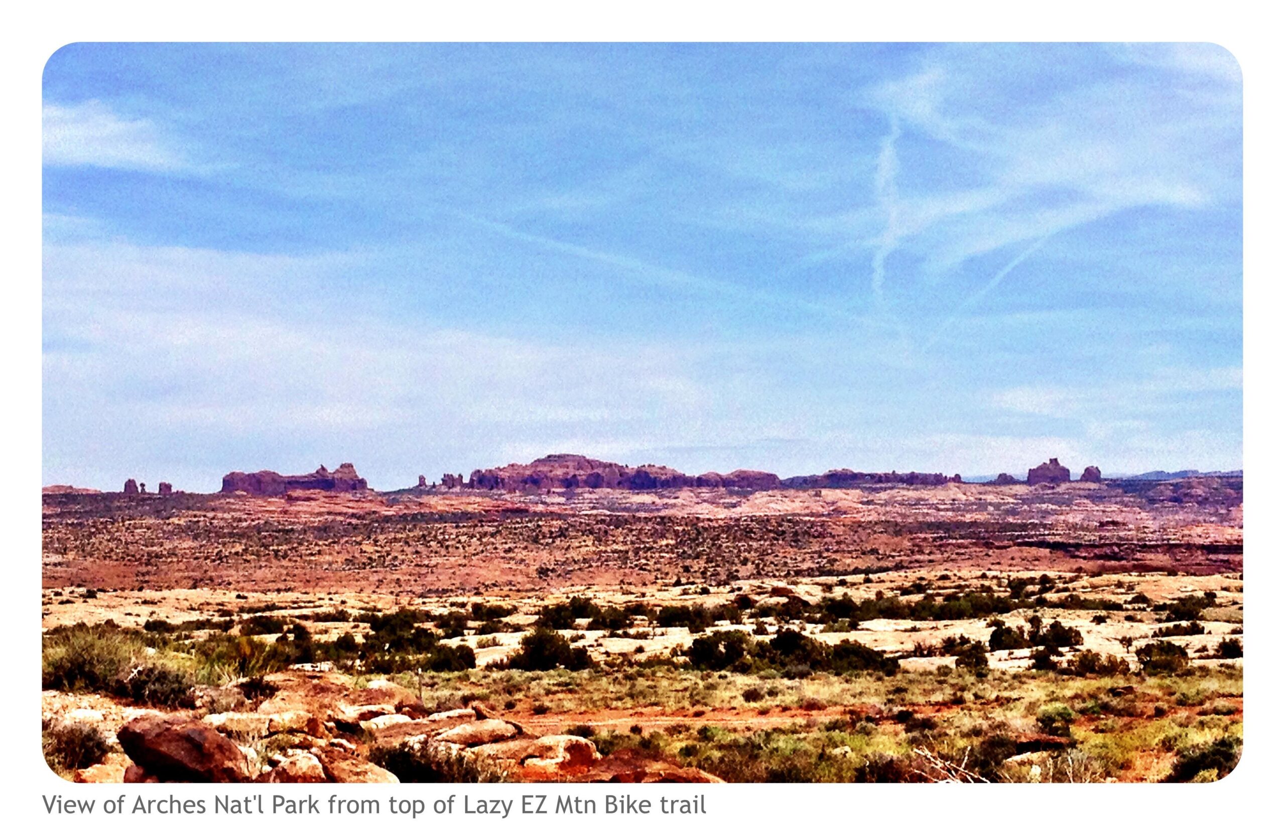 A scenic view of Arches National Park, captured from the top of the Lazy EZ Mountain Bike trail. The landscape features vast desert terrain with unique rock formations under a clear blue sky, showcasing the natural beauty of the park. Moab Brand Trails mountain bike trail.