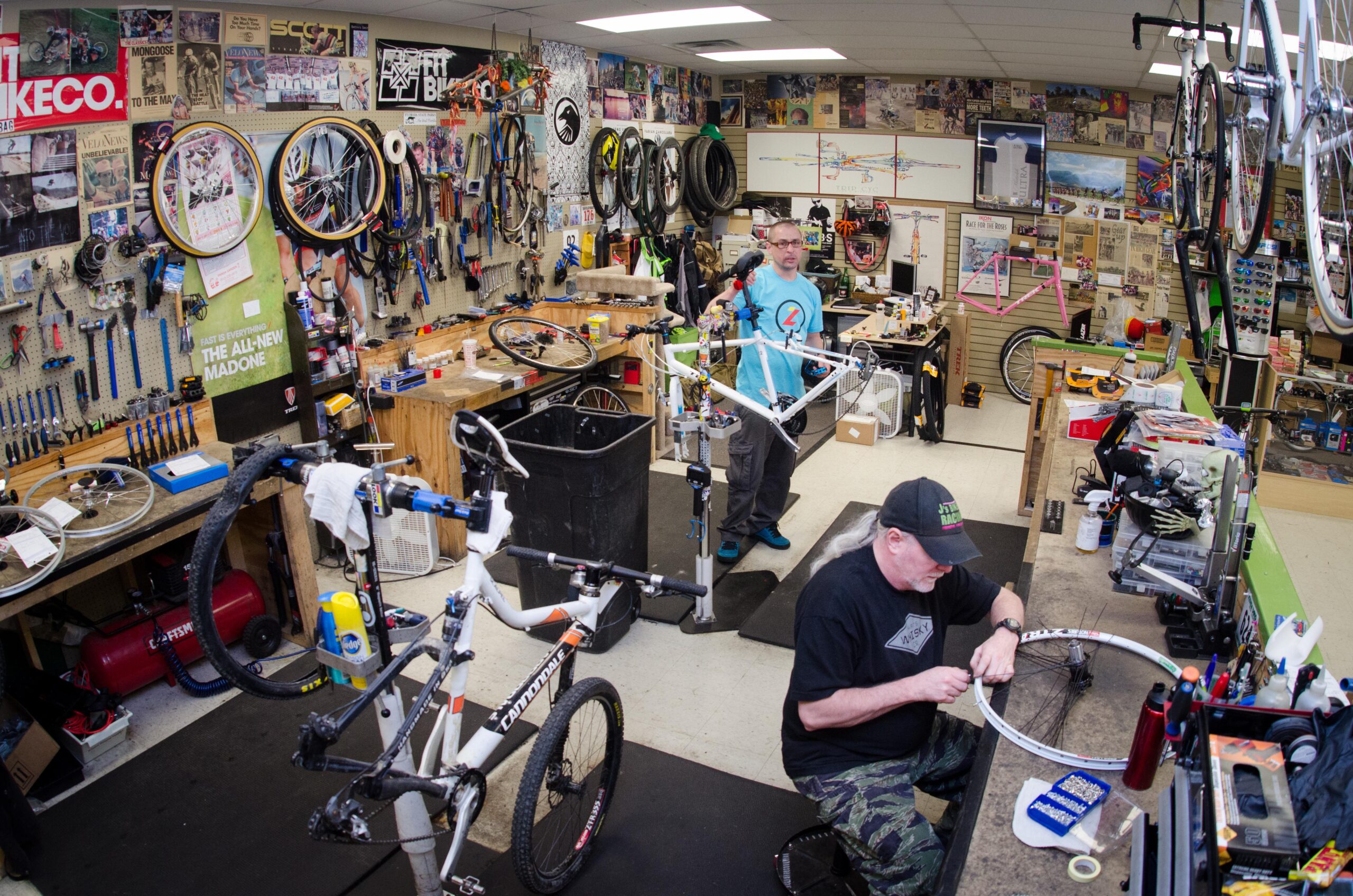 A busy bike repair shop featuring two mechanics working on bicycles. One mechanic stands with a partially assembled bike, while the other is seated at a workbench, adjusting a bike wheel. The walls are lined with tools, bike parts, and various cycling memorabilia, creating a vibrant and dynamic atmosphere.