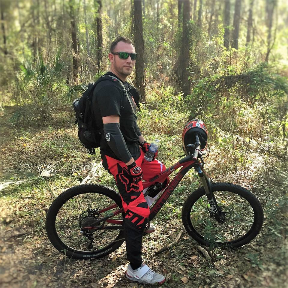 Intense Tracer 275: A man standing next to a mountain bike in a wooded area. He is wearing sunglasses, a black t-shirt, red cycling pants, and protective gloves. He has a backpack and is holding a water bottle. The background features trees and foliage, indicating a natural outdoor setting.