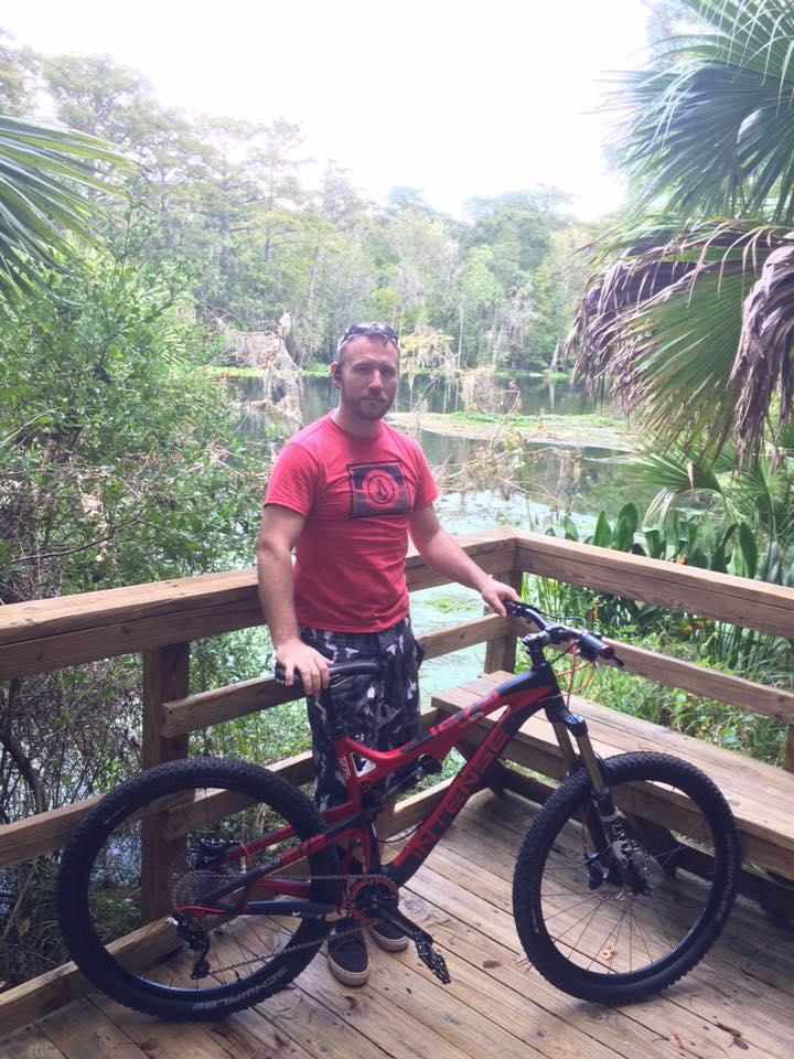 Intense Tracer 275: A man stands on a wooden deck beside a mountain bike, with a scenic view of a lake surrounded by trees and lush greenery in the background. He is wearing a red t-shirt and patterned shorts. The atmosphere appears peaceful and natural.