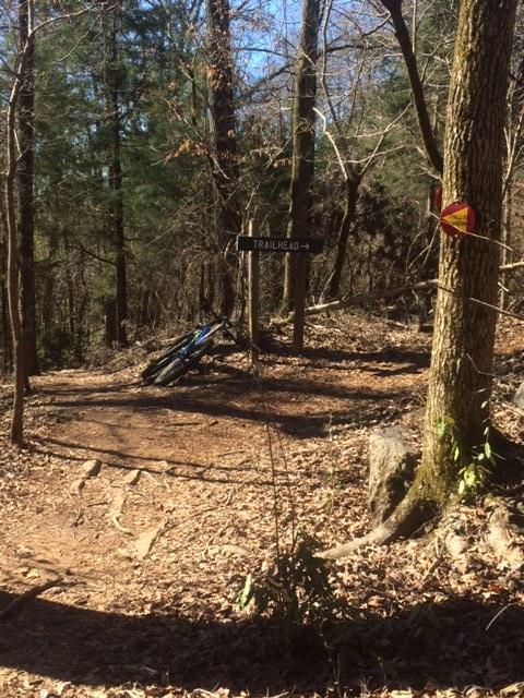 A winding dirt path leads to a sign labeled "TRAILHEAD" amidst a forested area with leafless trees. A mountain bike leans against the sign, and colorful trail markers can be seen on nearby trees. The ground is covered in fallen leaves and natural debris, indicating a serene outdoor environment. Chewacla State Park mountain bike trail.