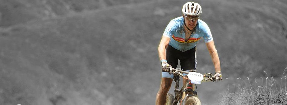 A mountain biker in a blue jersey and white helmet rides on a dirt trail, focused and determined. The background features a blurred, hilly landscape, emphasizing the athlete's movement and effort in the sport.