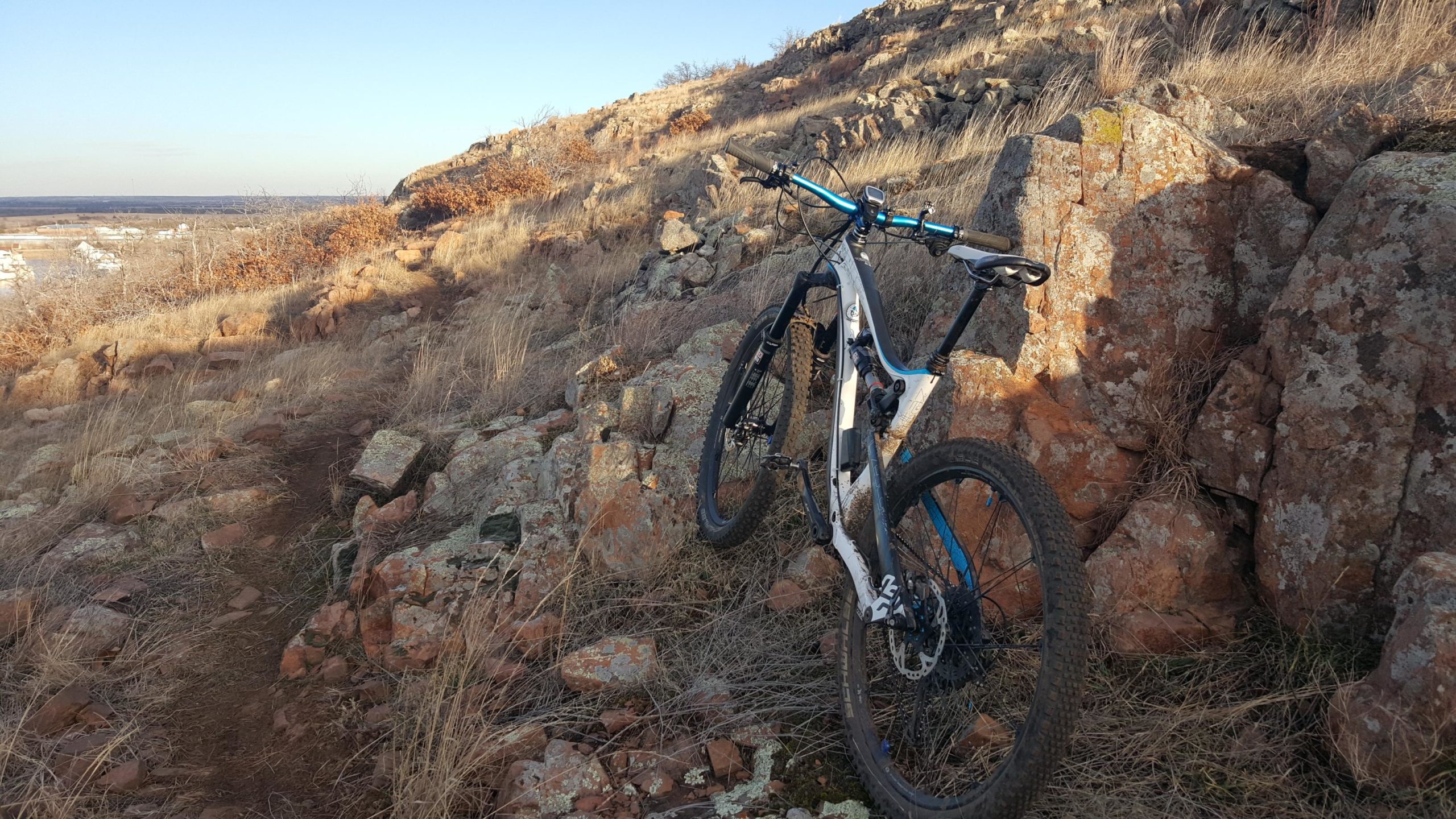 A mountain bike leaning against a rocky outcrop on a rugged trail, surrounded by dry grass and a scenic landscape in the background. The lighting suggests early morning or late afternoon, highlighting the texture of the rocks and foliage. Lake Lawtonka Trails mountain bike trail.