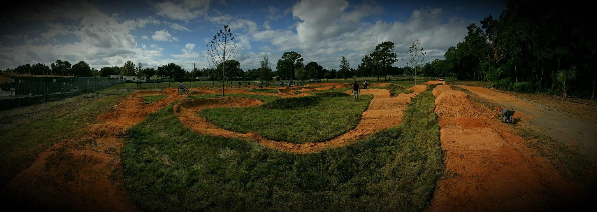 A panoramic view of a dirt bike park under construction, featuring winding dirt tracks surrounded by grassy areas. Several people are actively working on the site, with some riding bicycles along the paths. In the background, there are trees and a cloudy sky, contributing to the outdoor setting. A wheelbarrow is visible in the foreground, indicating ongoing earth-moving activities. Orlando Mountain Bike Park mountain bike trail.
