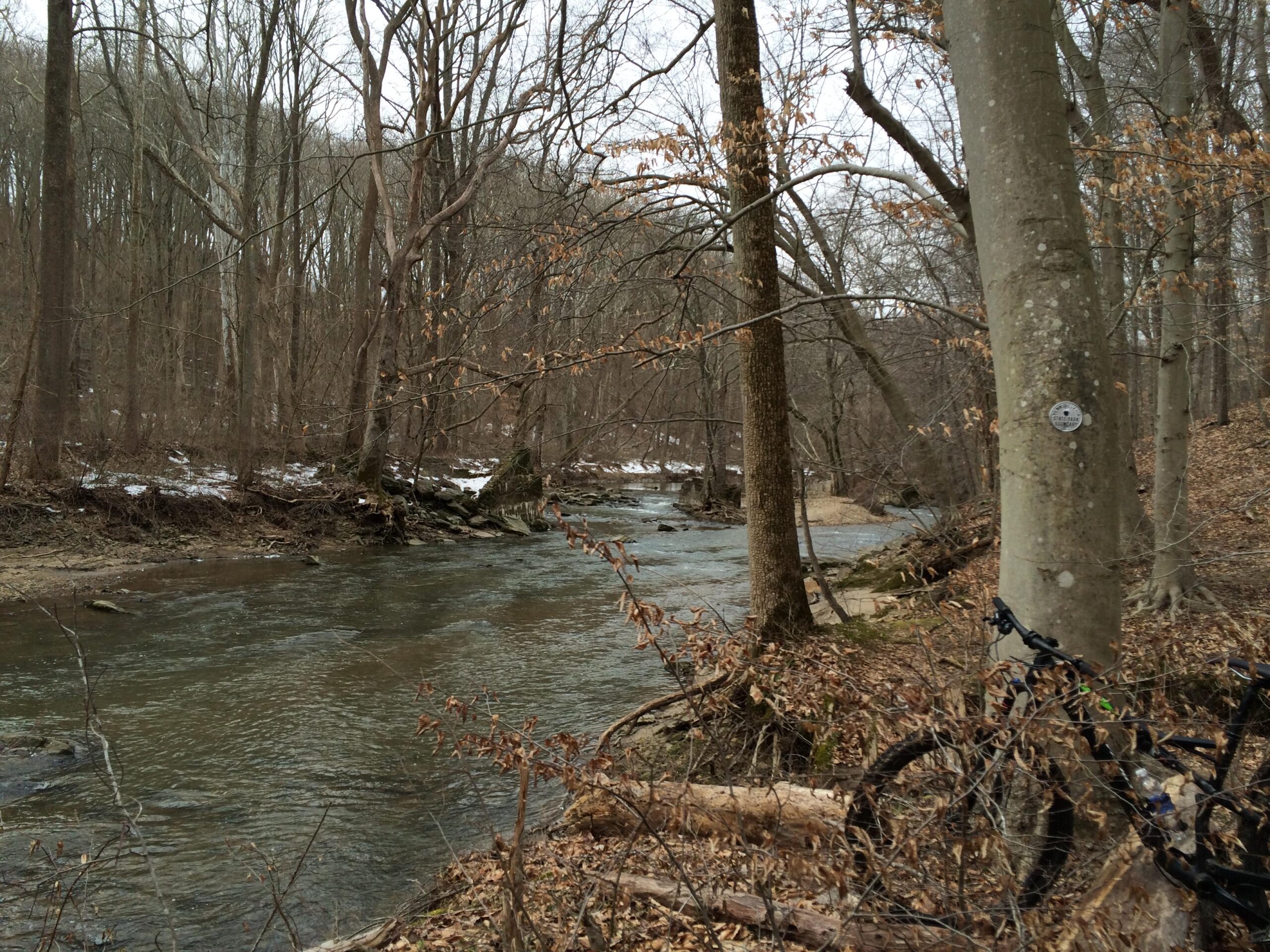 A tranquil stream flowing through a wooded area in early spring, with bare trees on either side. The water is clear, reflecting the surroundings, and some patches of snow can be seen on the ground. In the foreground, a bicycle is resting against a tree, and a small trail is visible along the bank. White Clay Creek mountain bike trail.