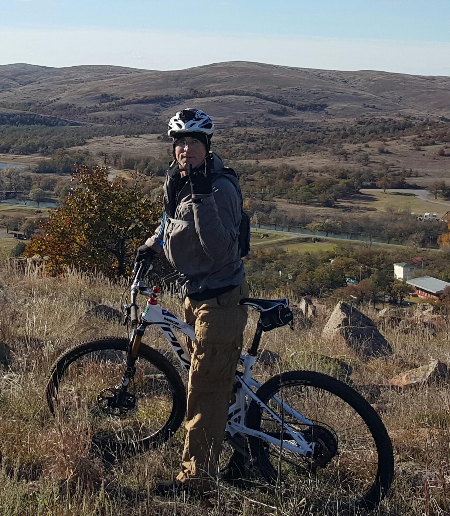 A person wearing a helmet and mountain biking gear stands next to a mountain bike, giving a hand gesture while overlooking a scenic landscape with rolling hills and trees in the background. The atmosphere is sunny, and the terrain is grassy with some rocks. Lake Lawtonka Trails mountain bike trail.