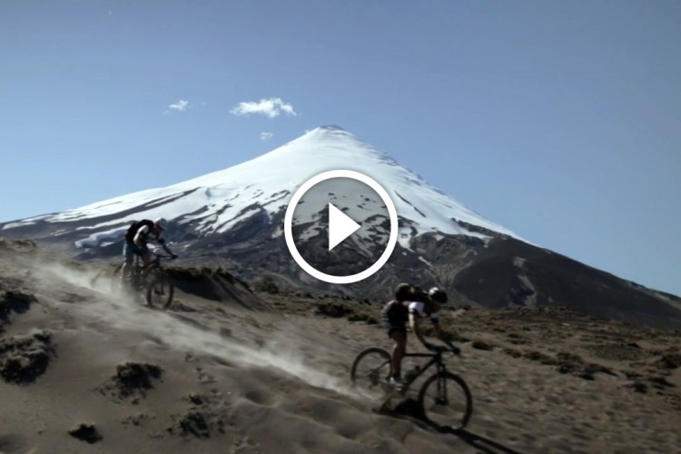Two mountain bikers riding on a dusty trail with a snow-capped volcano in the background. The sky is clear, and the scene captures a sense of adventure and outdoor activity.