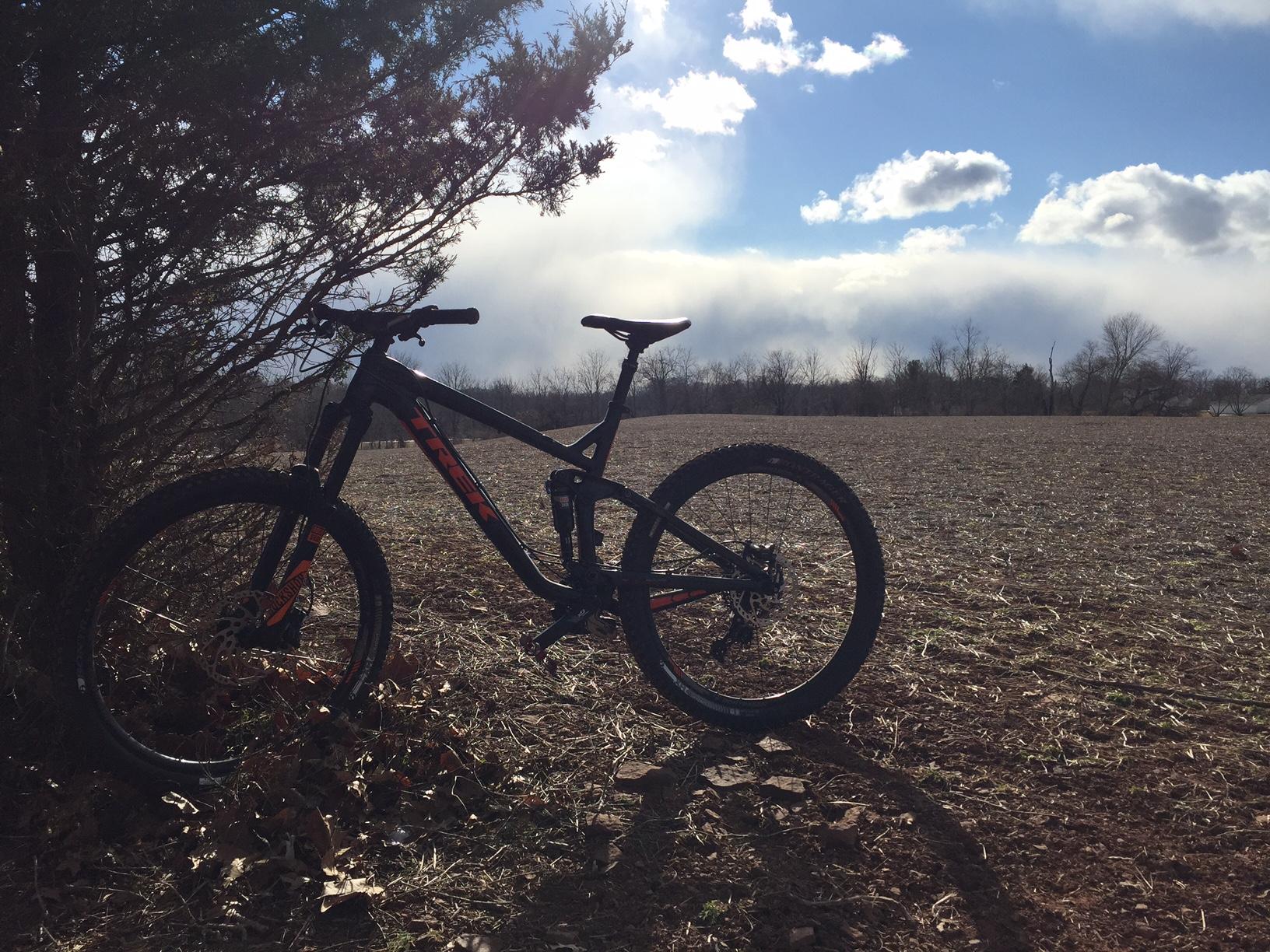 A black and orange mountain bike is leaning against a bush in an open field under a partly cloudy sky. The landscape is barren with some scattered leaves and faint outlines of trees in the background. Six Mile Run mountain bike trail.