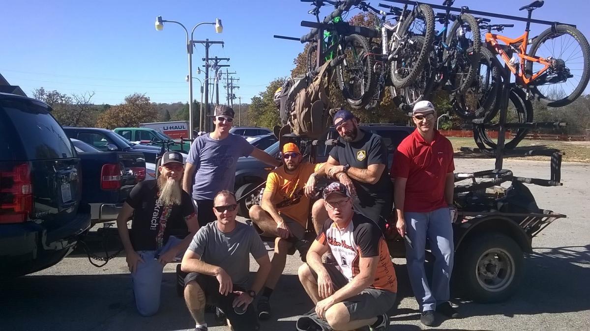 A group of seven men posing together in a parking area, with several mountain bikes mounted on a rack behind them. The men are dressed in casual clothing, including t-shirts and caps, and they appear to be enjoying a sunny day outdoors. The background shows vehicles and power lines, indicating a recreational setting.