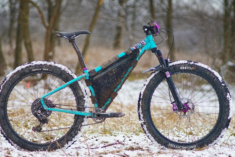 A mountain bike with a vibrant turquoise and black frame, equipped with wide tires partially covered in snow, stands in a winter landscape. The surrounding area features grass and trees, with light snow falling. The bike has a modern design, showcasing purple accents on the handlebars and wheels.