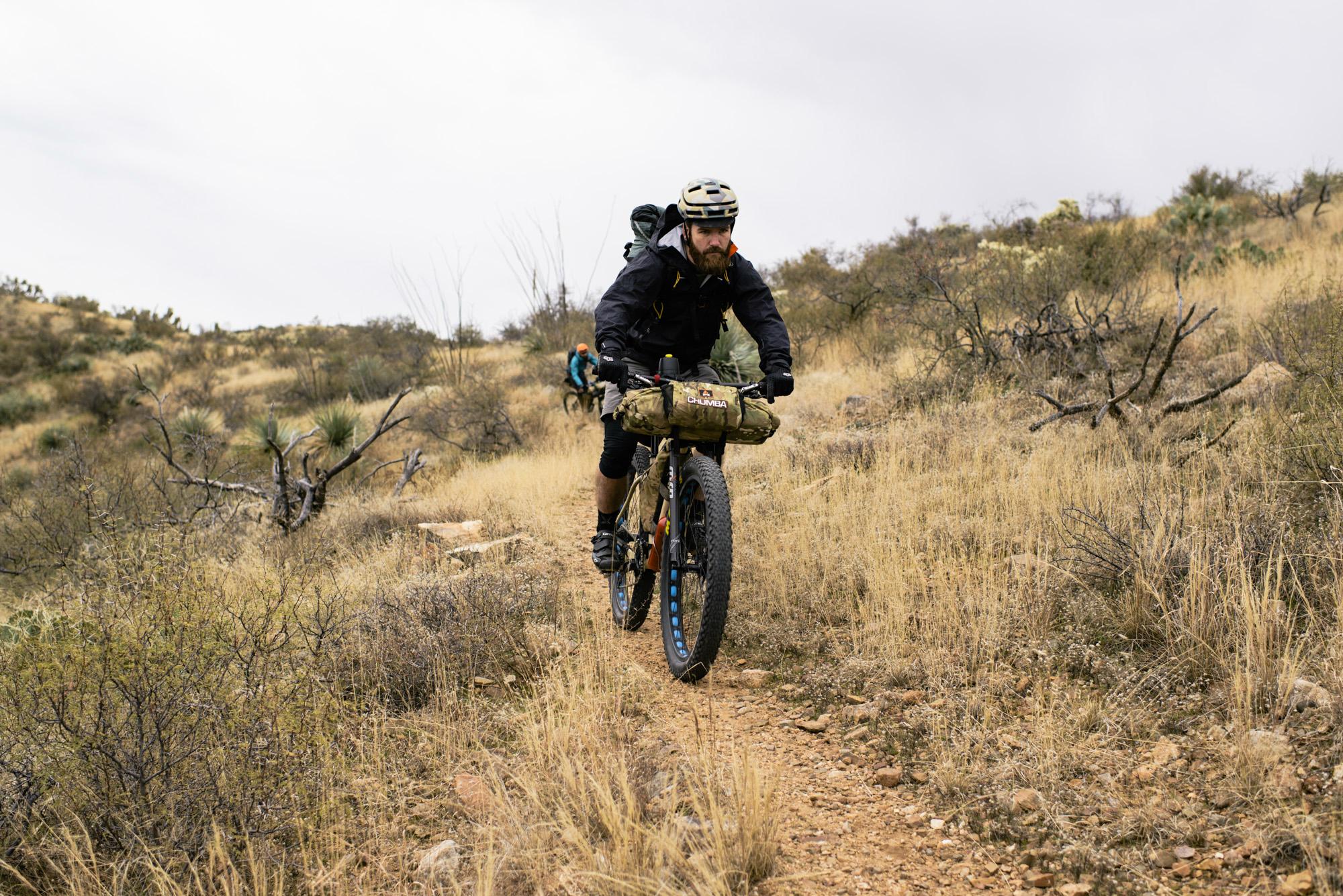 A cyclist navigating a dirt trail through a rugged landscape with dry grass and sparse vegetation. The rider is wearing a helmet and cycling gear, and is carrying gear on the bike. In the background, another cyclist can be seen on the trail. The sky is cloudy, indicating overcast weather. Arizona Trail mountain bike trail.