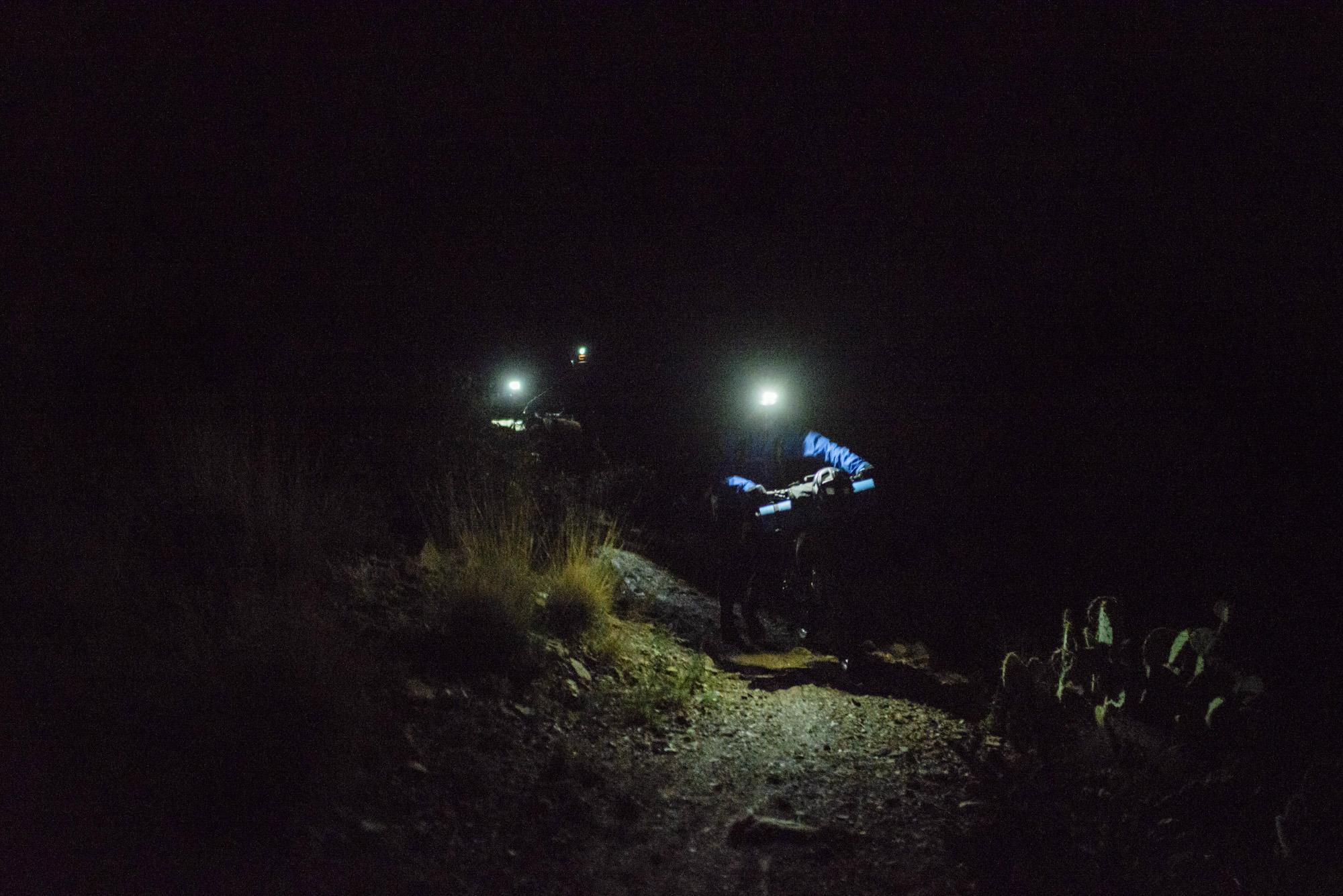 Two hikers ascend a narrow, rocky path at night, illuminated by headlamps. The scene is dark, with only the beams of light highlighting the trail and surrounding vegetation. Sagebrush and cacti are visible along the path. Arizona Trail mountain bike trail.