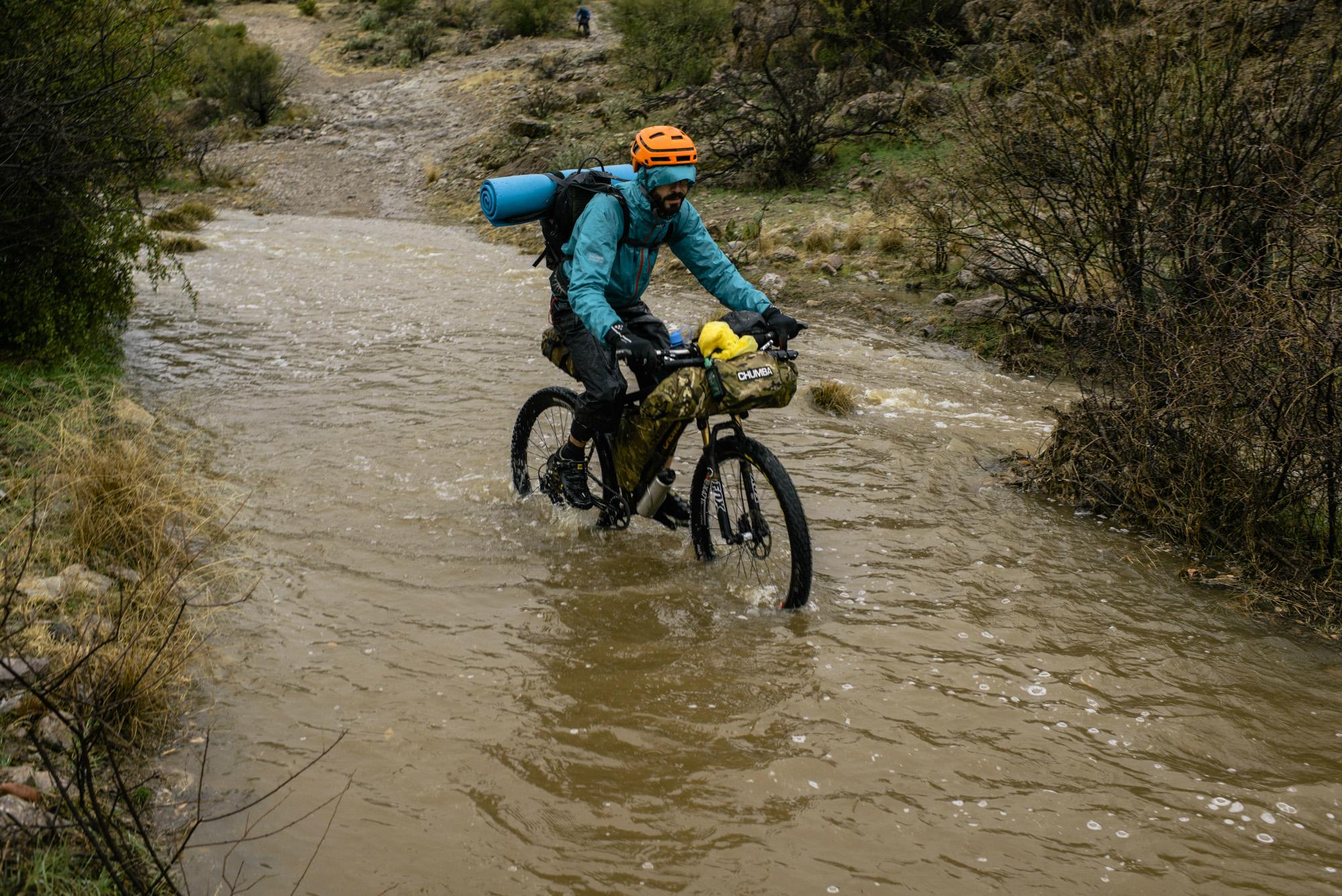 A cyclist wearing a helmet and a waterproof jacket navigates through a flooded dirt path on a mountain bike. The bike is equipped with bags and a blue sleeping pad is secured on top. The surroundings are a mix of wet terrain and sparse vegetation. Arizona Trail mountain bike trail.