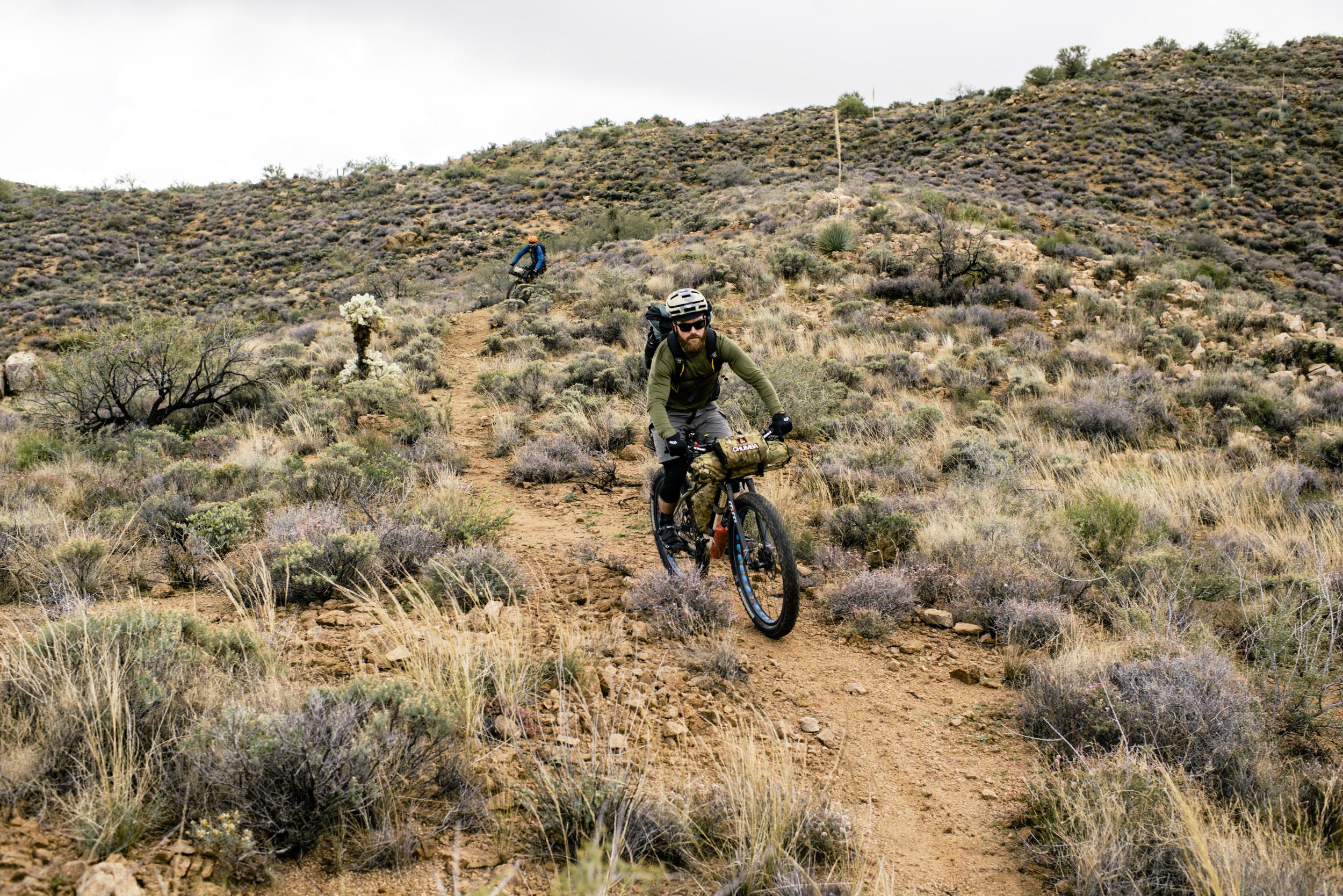 Two mountain bikers navigating a rugged trail in a hilly, desert landscape filled with sparse vegetation and dry grasses. One rider, wearing a helmet and sunglasses, is cycling towards the viewer, while another rider is seen further along the path in the background. The atmosphere is overcast, suggesting a cooler day for biking. Arizona Trail mountain bike trail.